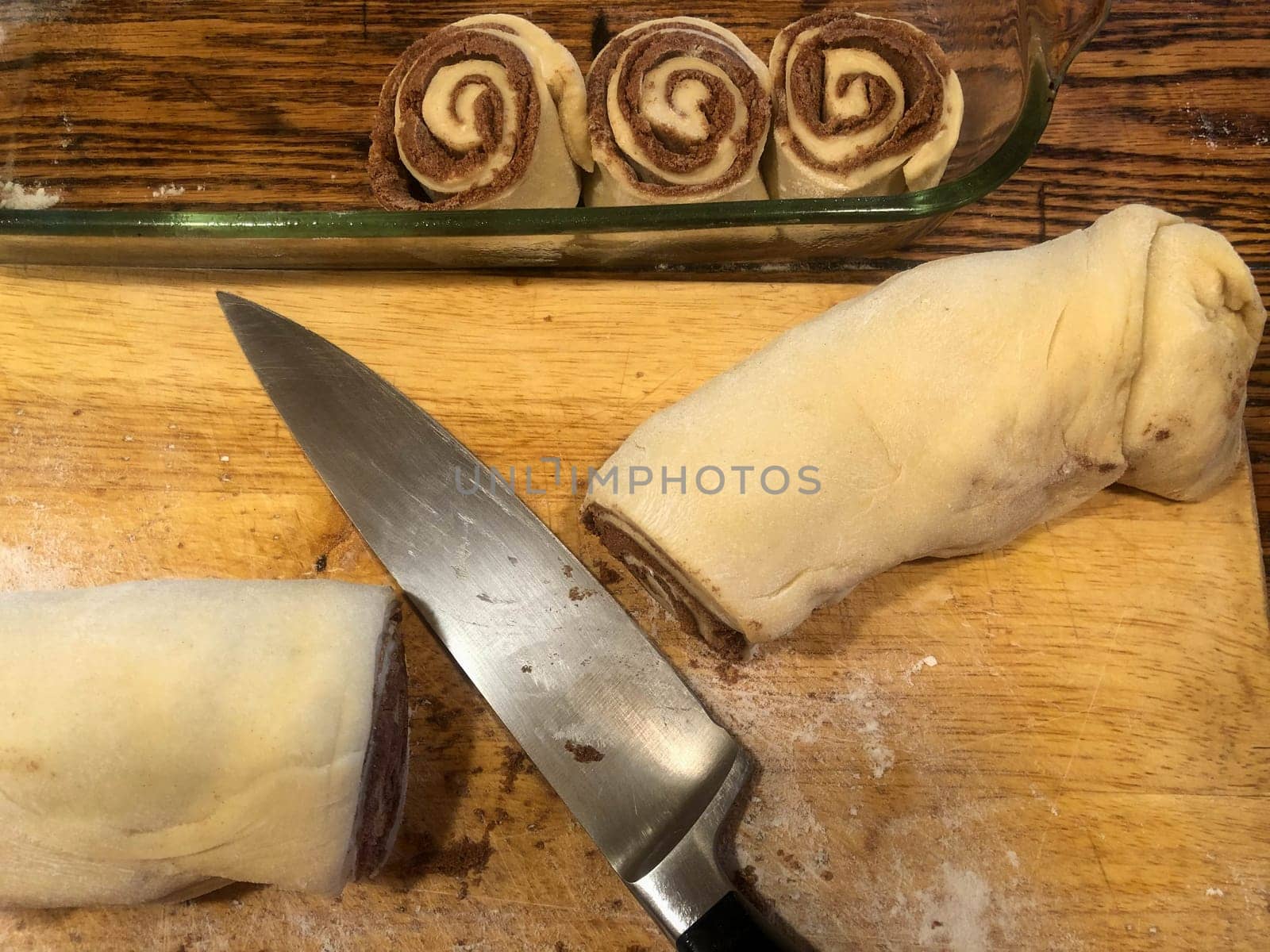 Beautiful hand rolled cinnamon buns with knife and cut rolls in baking dish. High angle view of rustic home bakery natural look with quality steel knife blade, glass dish, and wooden cutting board