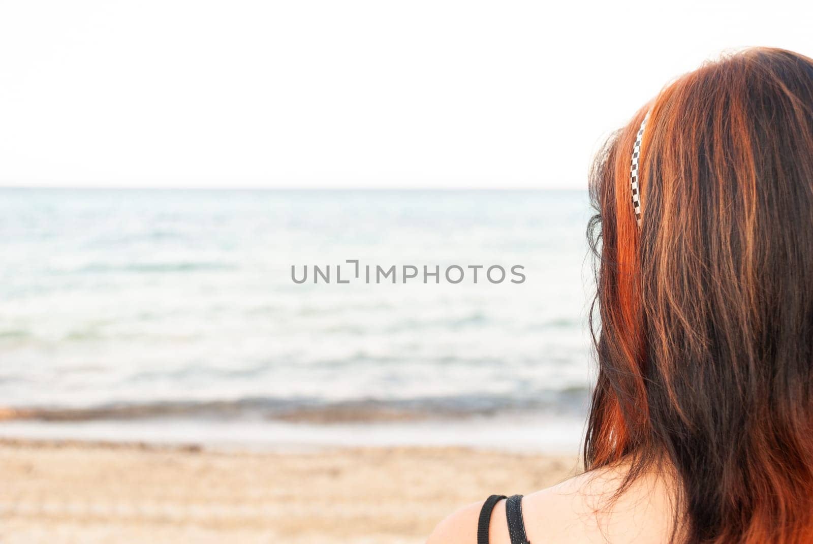 Young woman with red and black long hair standing at the beach, looking at the sea, rear view, horizontal