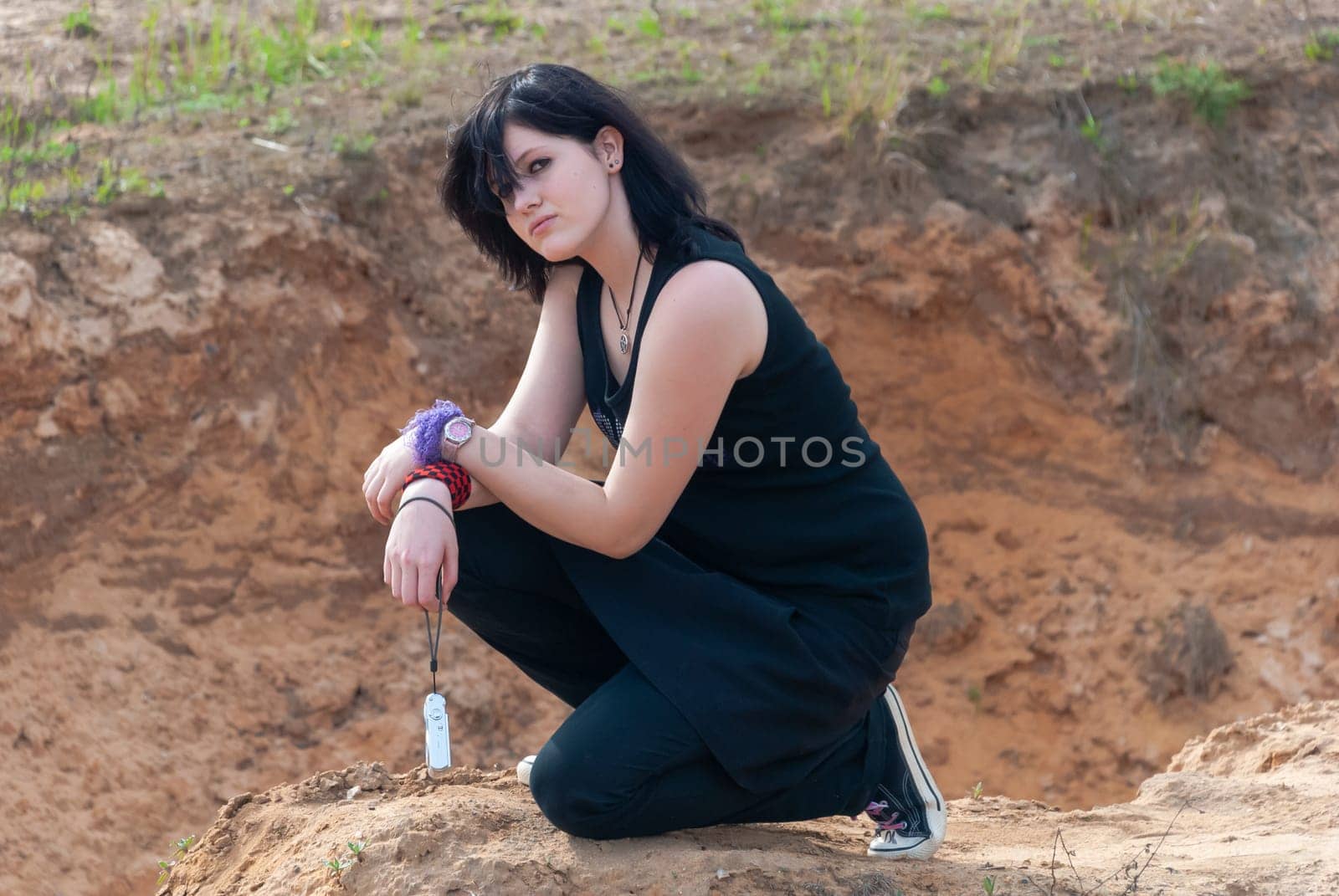 Punk emo girl, young adult with long black hair, kneeling on a hill outdoors in the nature, holding a camera in her left hand, looking at camera, horizontal