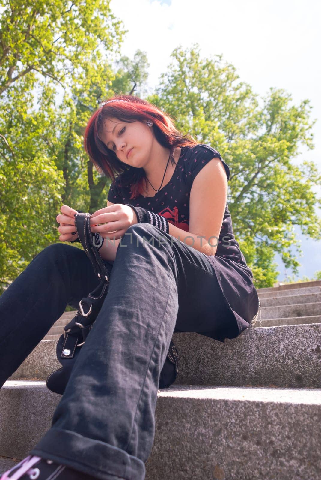 Punk emo girl, young adult with black red hair, sitting on a staircase outdoors, looking down, low angle view, vertical
