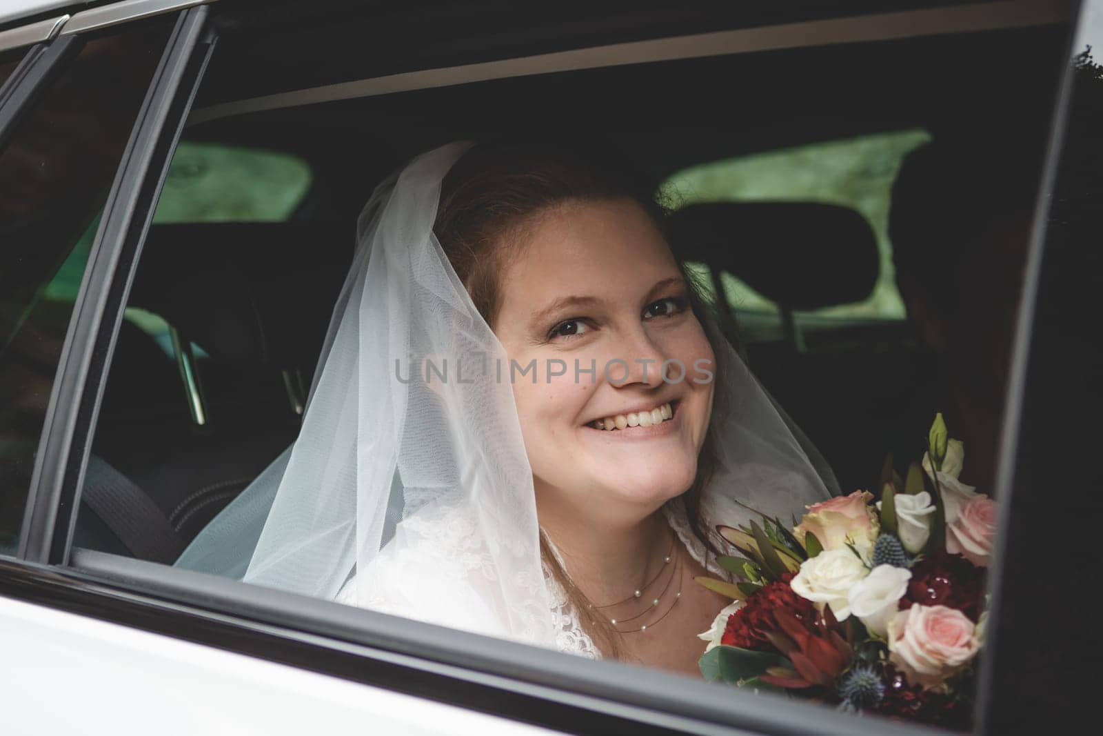 Bride with brown hair, sitting in car on her way to her wedding, holding flowers, smiling and looking at camera through a car window