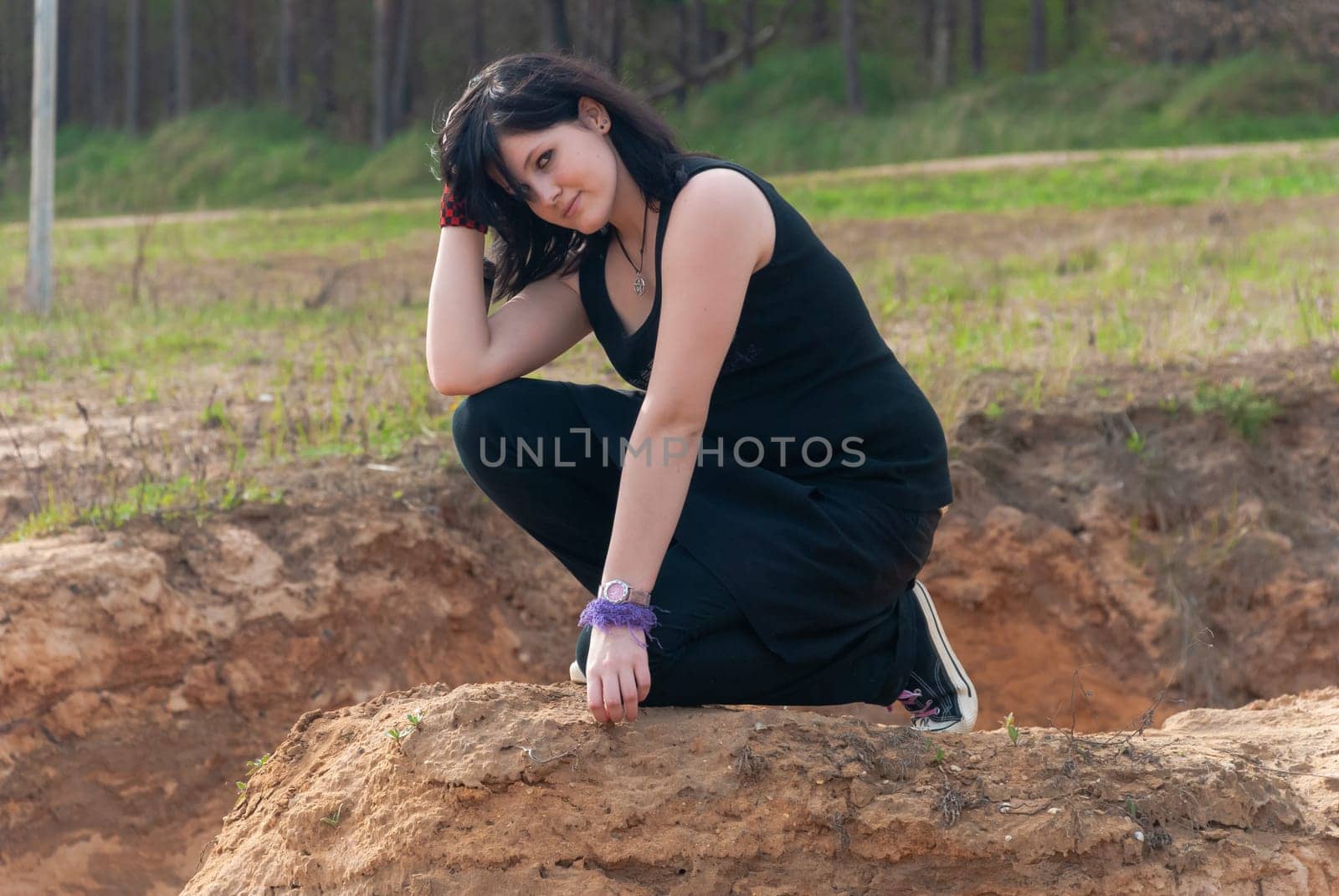 Punk emo girl, young adult with long black hair, kneeling on a hill outdoors in the nature, looking at camera, horizontal