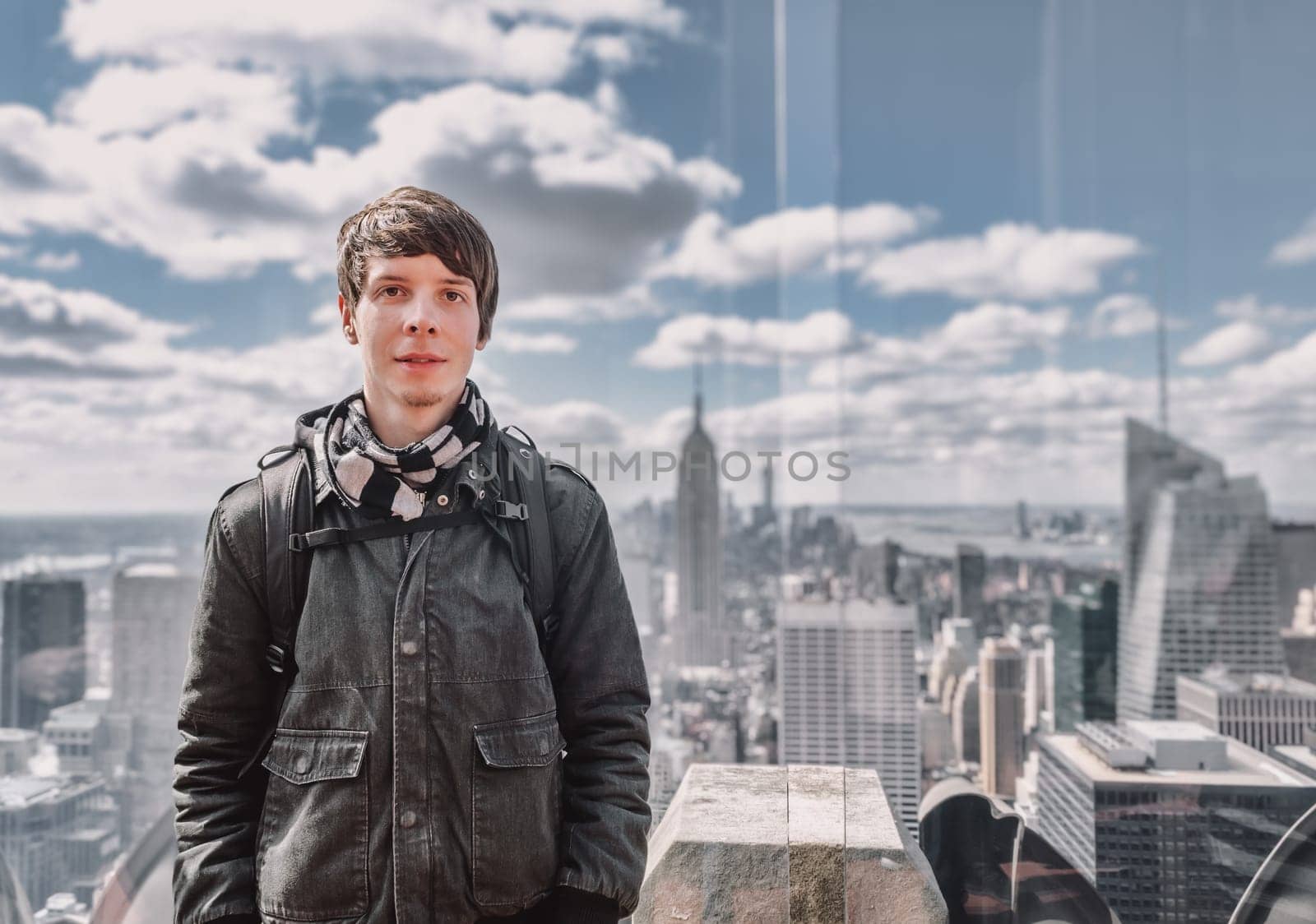 Young adult man with brown hair and beard looking at camera while standing on a observation tower in front of new york city skyline, usa, america