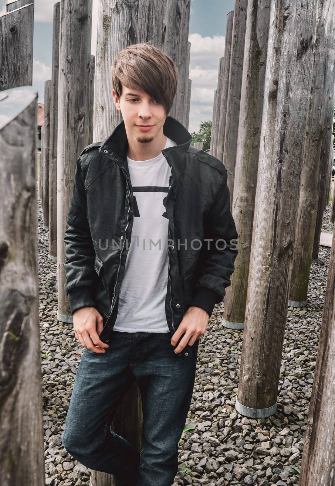 Young adult rock musician, singer wearing a black jacket and t-shirt leaning against wooden column looking down, brown hair and beard