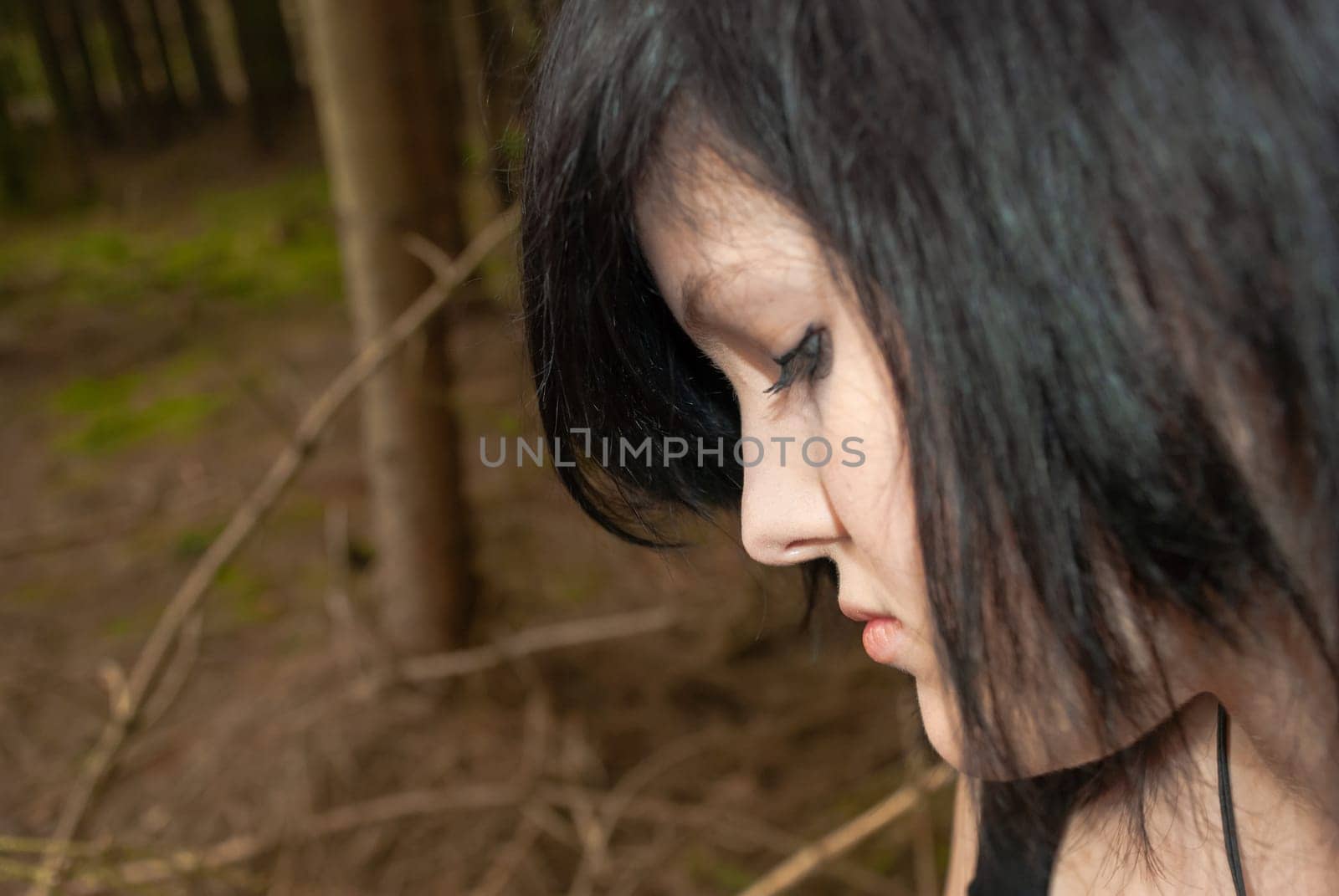 Punk emo girl, young adult with black hair and eyeliner, looking down, forest, close-up, horizontal