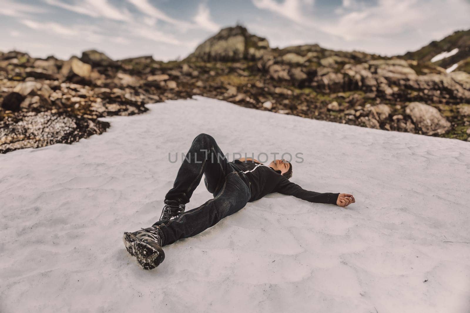 Young adult man is exhausted and relaxes after hiking to the top of a mountain in norway, lying down in the snow putting his hand on his breast