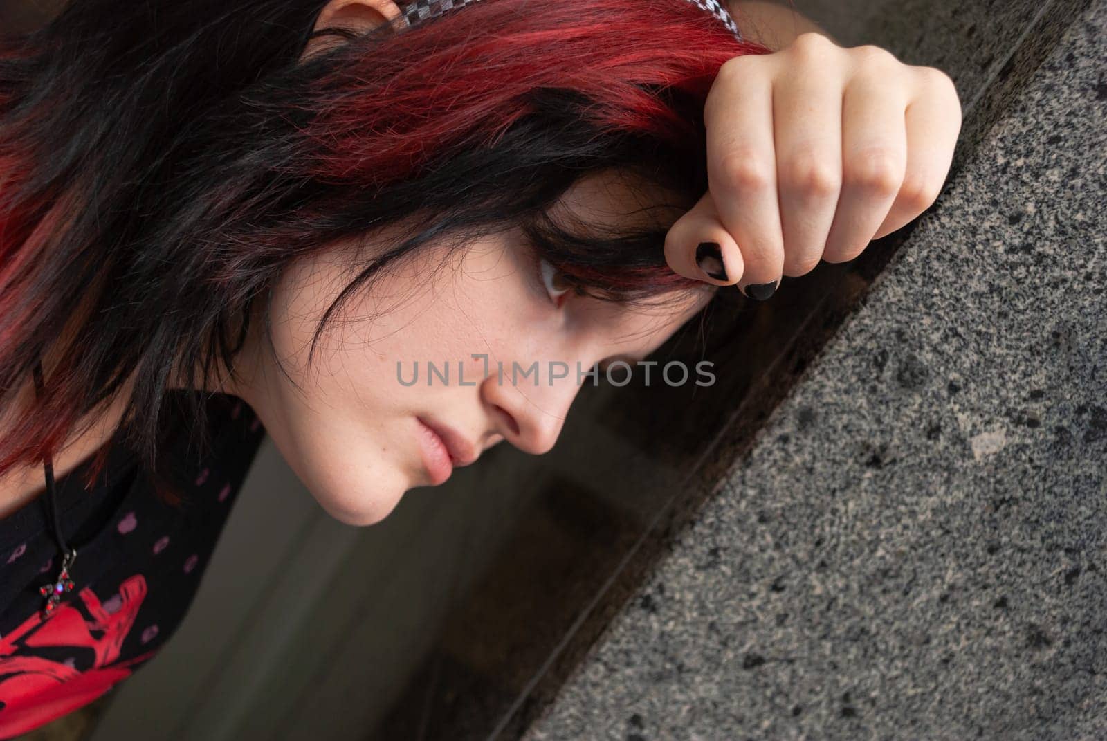Punk emo girl, young adult with black red hair, holindg her head against a concrete wall, close-up, horizontal