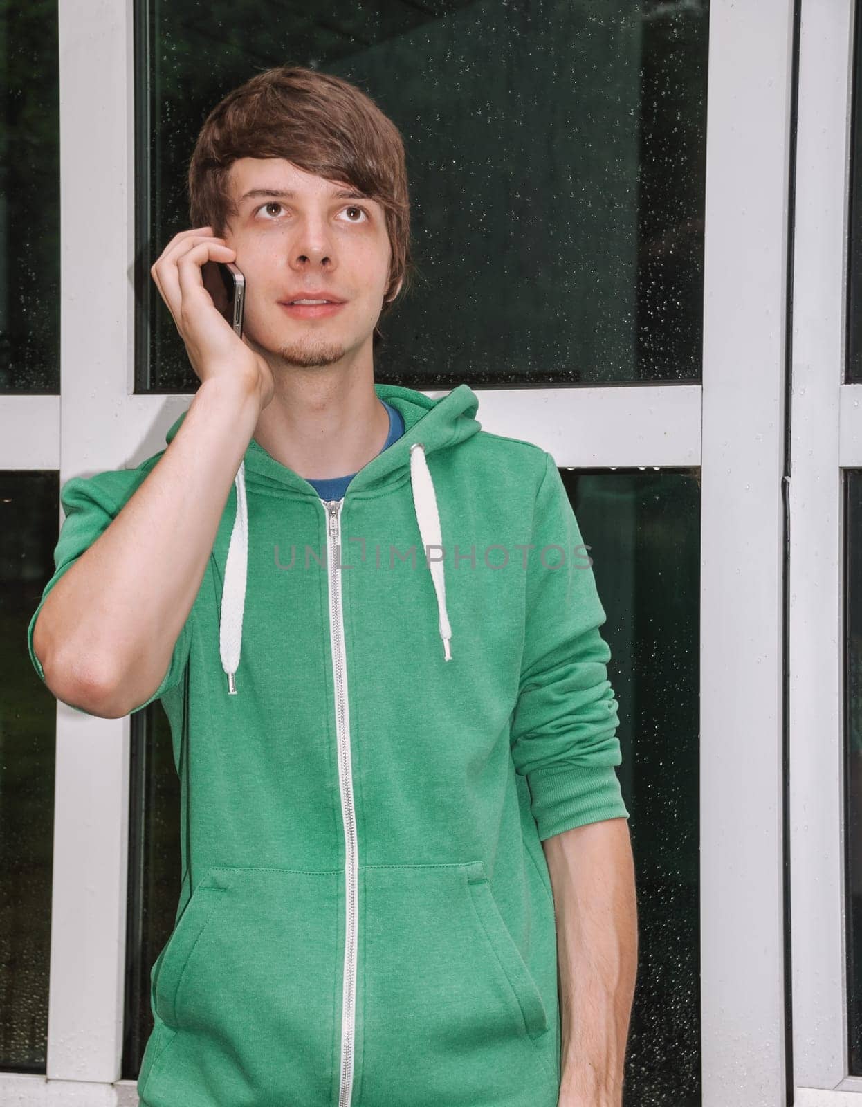 young man with brown hair and a beard is talking on the phone with his smartphone in front of his door with raindrops, wearing a green sweater, vertical