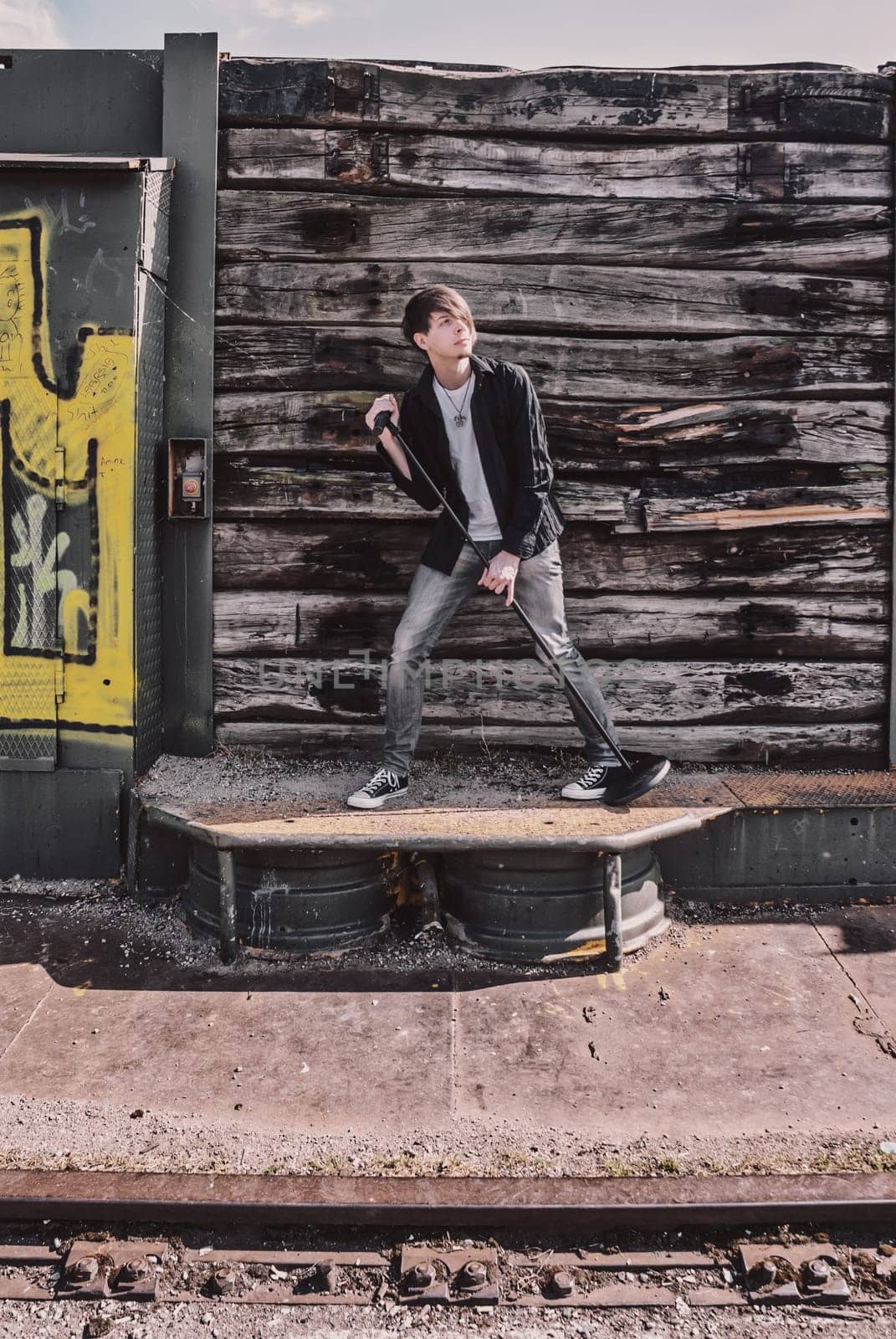 Young adult singer, with brown long hair and beard performing, rock musician with microphone outdoors in front of a wooden wall, standing, looking away, wearing jeans and t-shirt