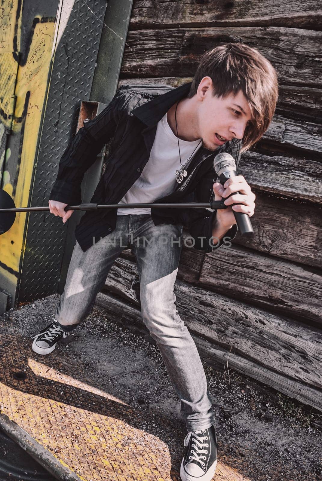 Young adult singer, with brown long hair and beard performing, rock musician with microphone outdoors in front of a wooden wall, standing, looking away, wearing jeans and t-shirt