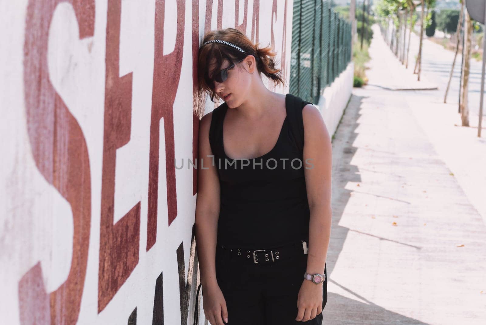 Young woman with black orange hair, wearing sunglasses and black casual clothing, leaning exhausted against wall to take a break from a long hike, front view, horizontal