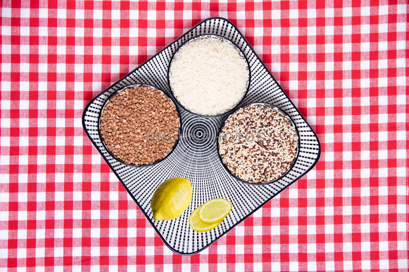 Vegetable food. Close-up of a decorative plate with three bowls with buckwheat, rice, quinoa and lemon on a red checkered tablecloth. Top view with space for design.