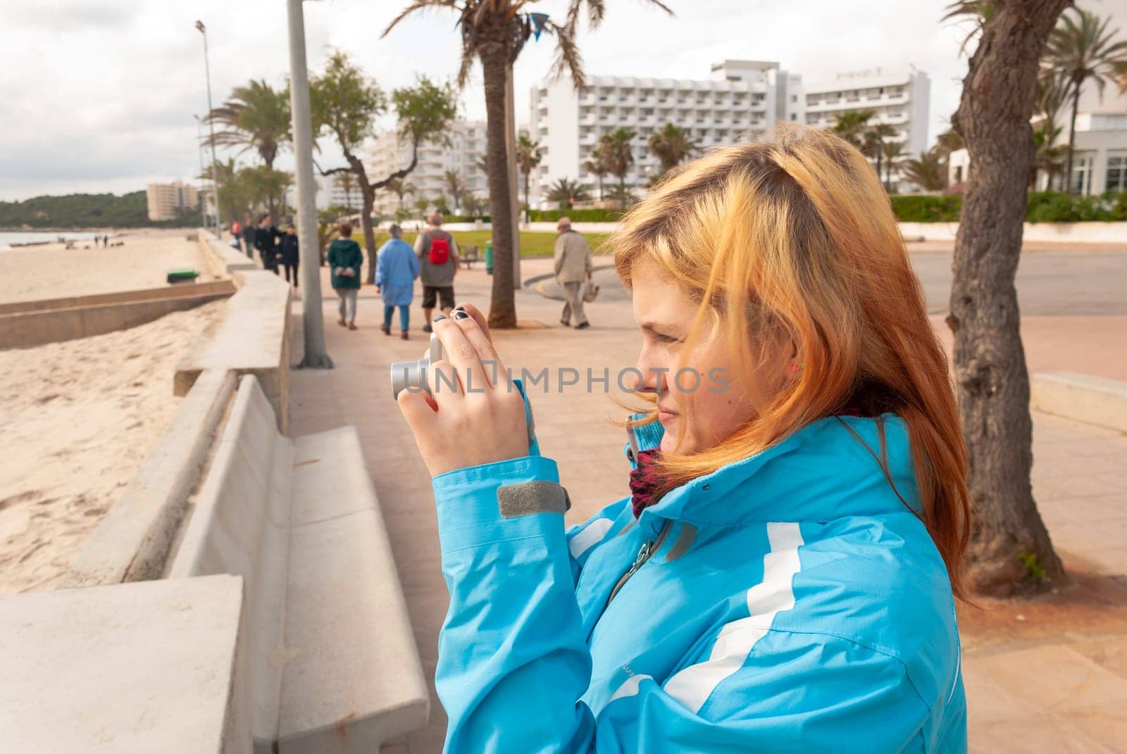 Young woman with blond hair, wearing a blue jacket standing at the promenade, taking photos of the beach in Cala Millor, Majorca