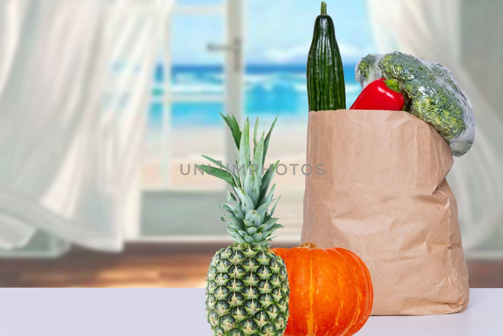Close up of a brown paper shopping bag with fresh vegetables, a pumpkin and a pineapple on the table over an abstract blurred sea background. Concept of strengthening the immune system. Copy space.