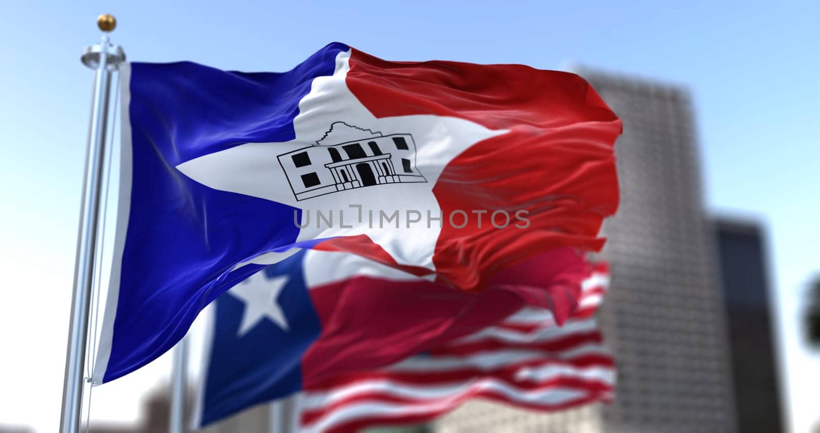 San Antonio city flag waving in the wind with Texas state and United States national flags blurred in background. Black-outlined Alamo in the middle of the flag