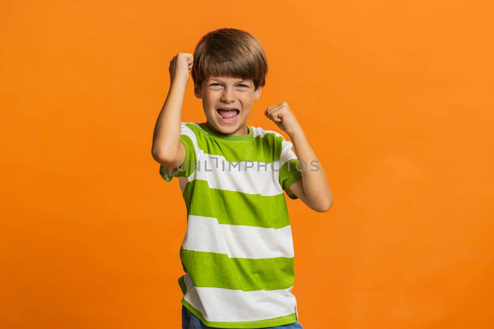 Young preteen child boy kid with crossed fingers wishing luck shouting, raising fists in gesture I did it, celebrating success, winning, birthday, lottery jackpot game achievement on orange background