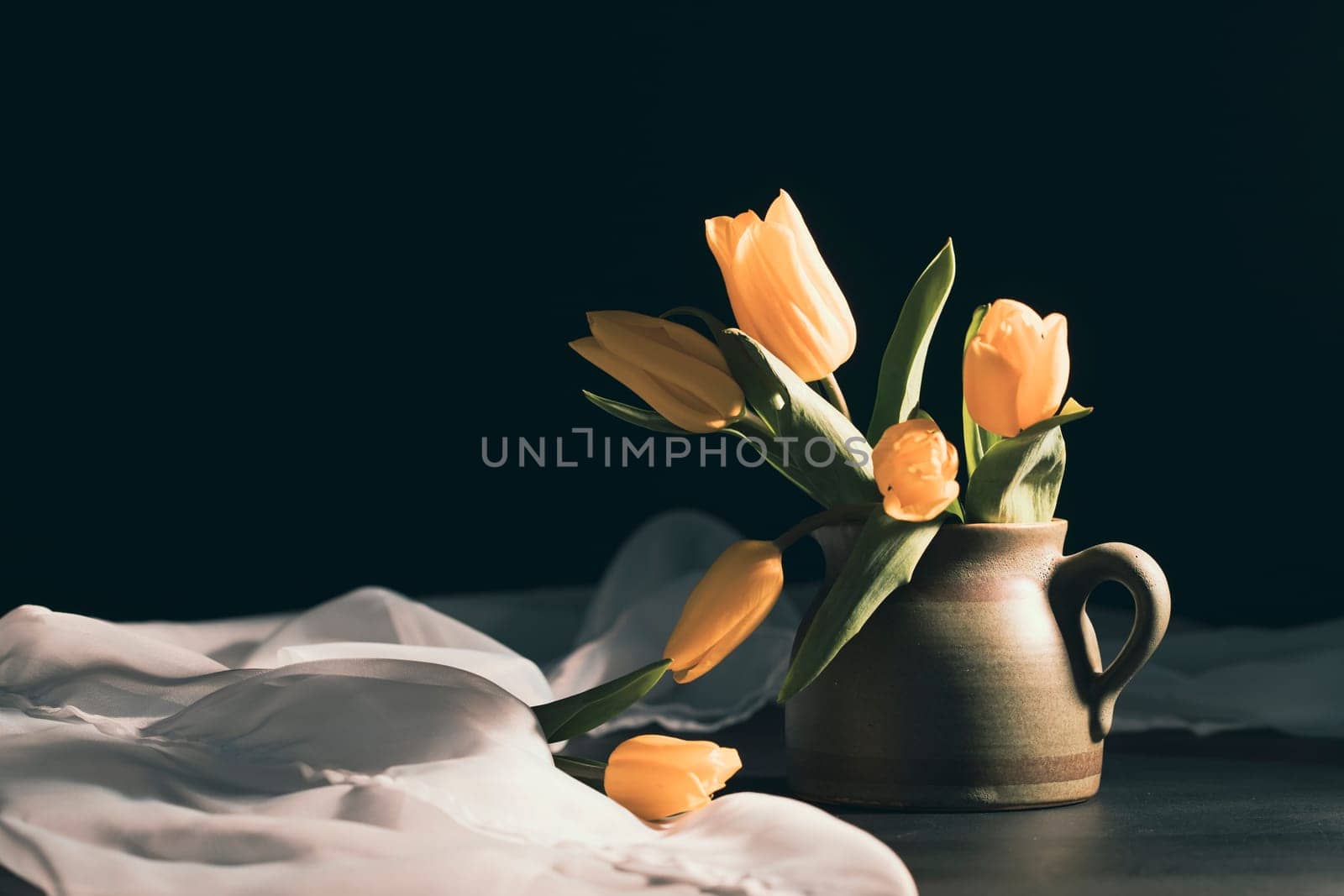 Still life with bouquet of yellow tulip flowers in old vintage jug, in beam of light on dark background.Art photography.