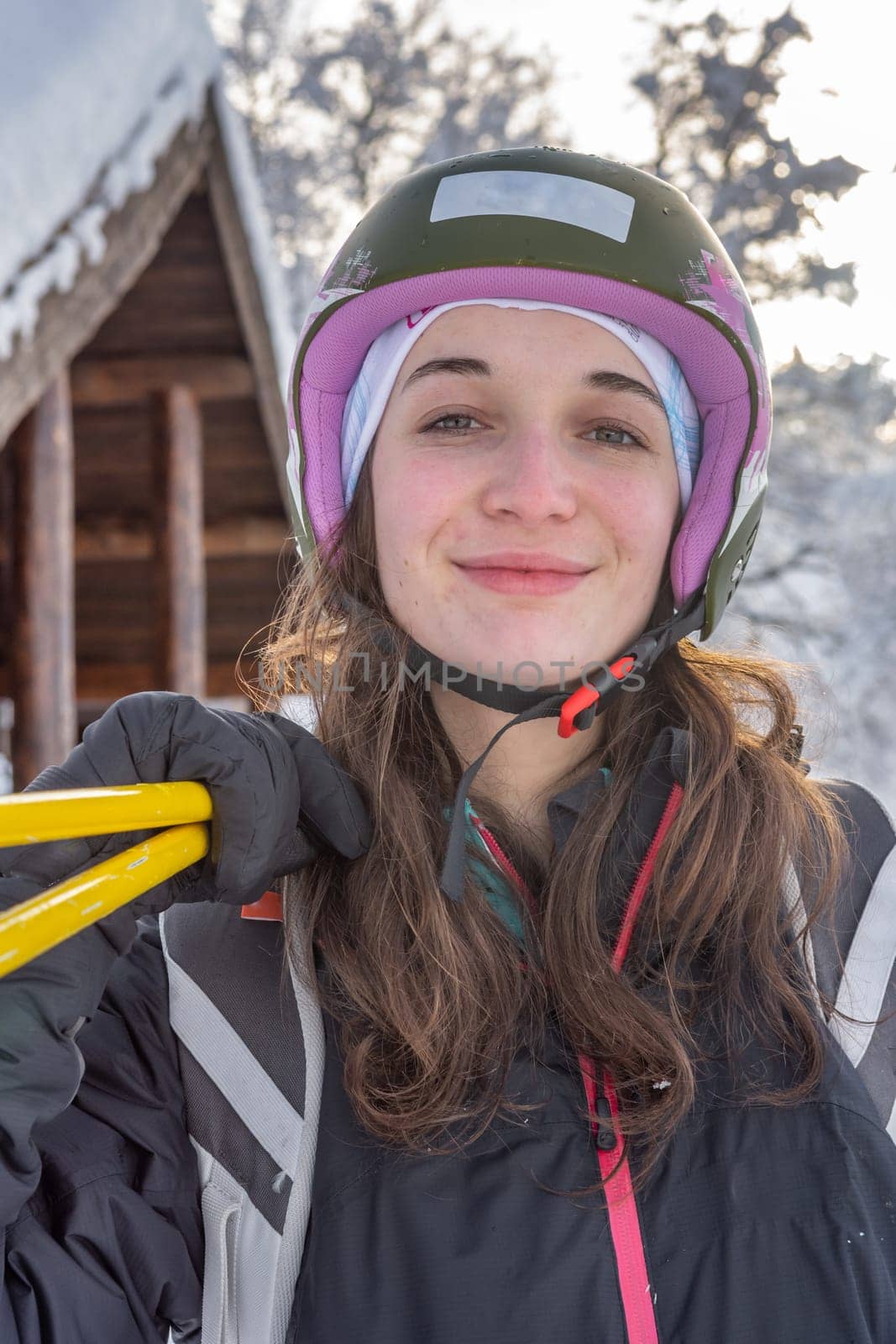 Woman with brown hair smiling, looking at camera, wearing ski wear and a ski helmet, holding ski poles on her right shoulder, close-up, in  skiing stryn, norway