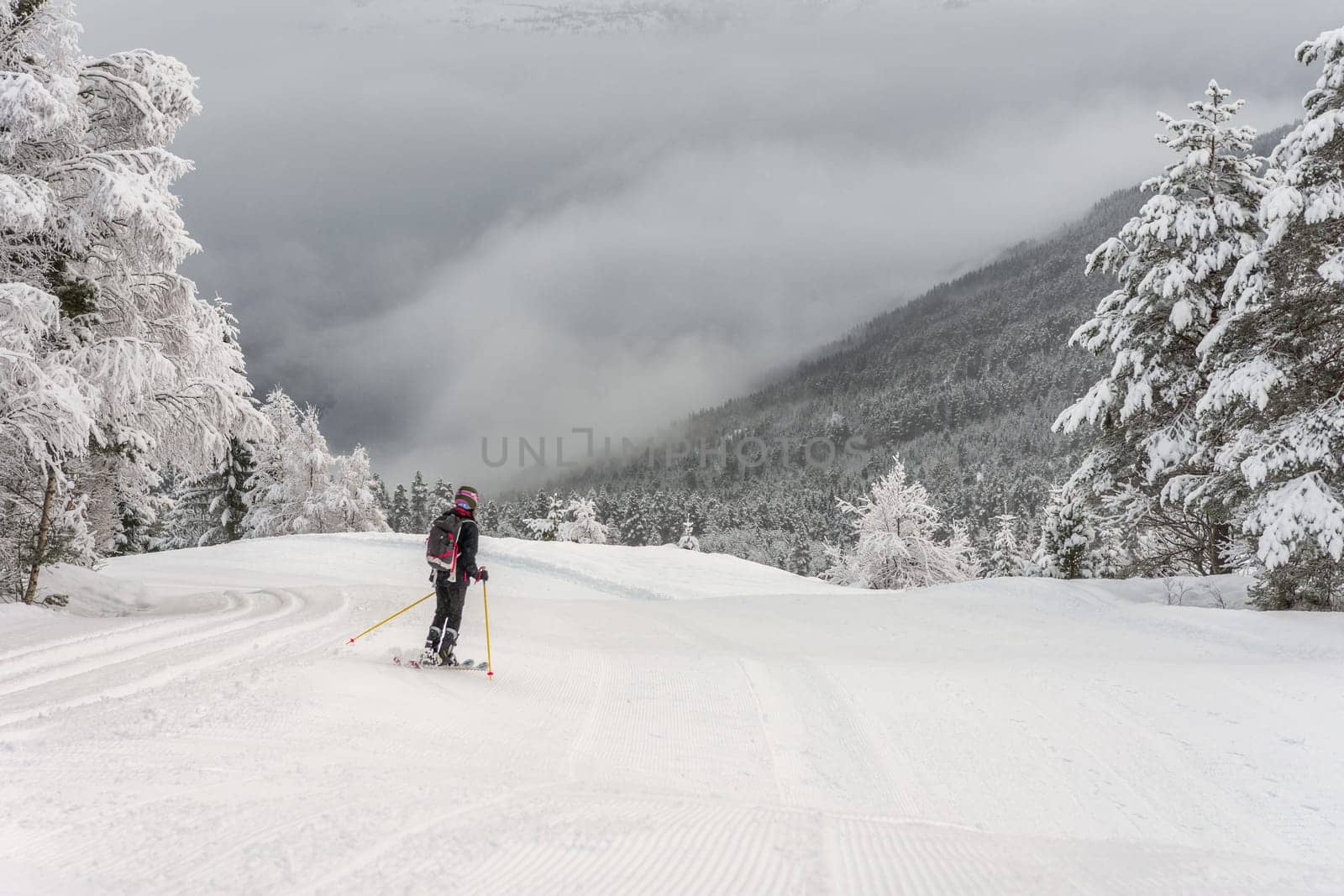 Woman with black ski wear, helmet and backpack skiing down the top of the mountain in ski centre, stryn with clouds and trees in the background, rear view, horizontal