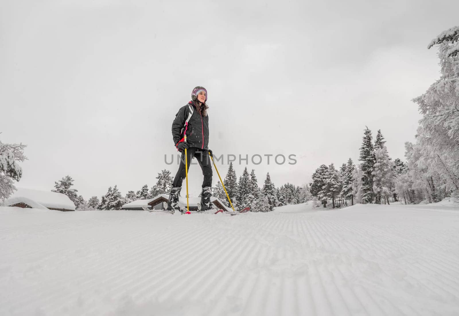Woman with brown hair, wearing ski wear, standing in the valley after skiing down the top of the mountain, overcast low angle view, horizontal