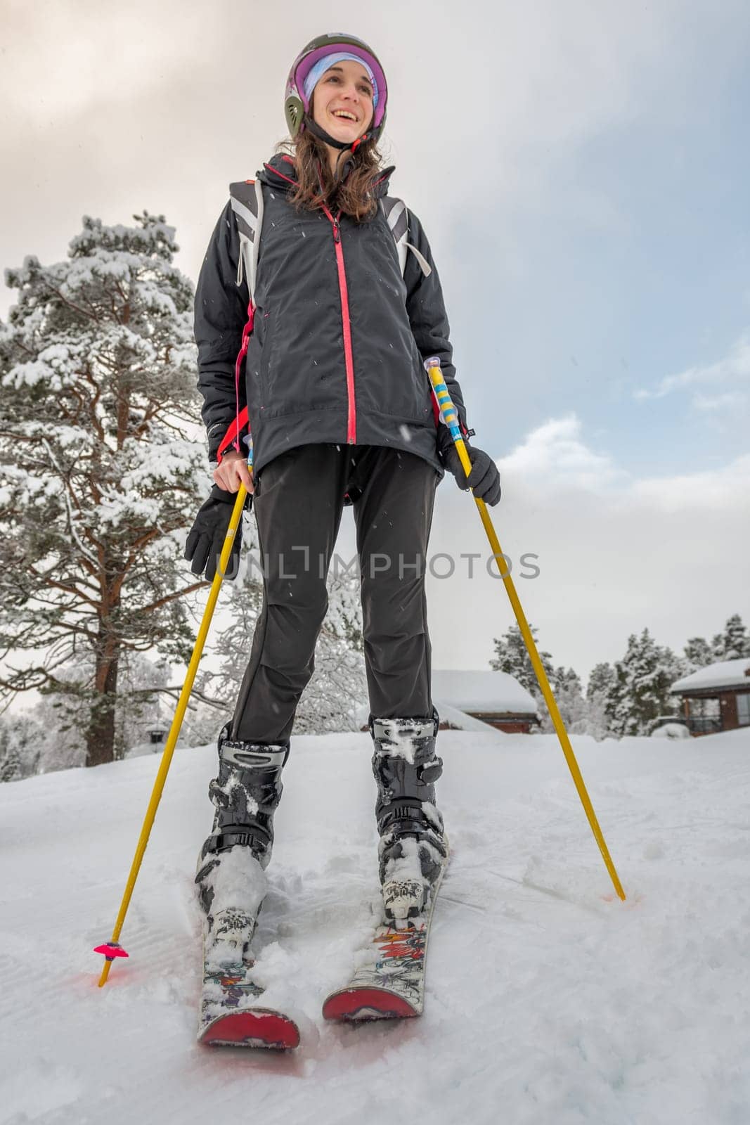 Ski Woman with brown hair, wearing black ski wear and helmet, holding yellow ski poles, smiling while standing on top of the mountain, looking at view, low angie view, vertical