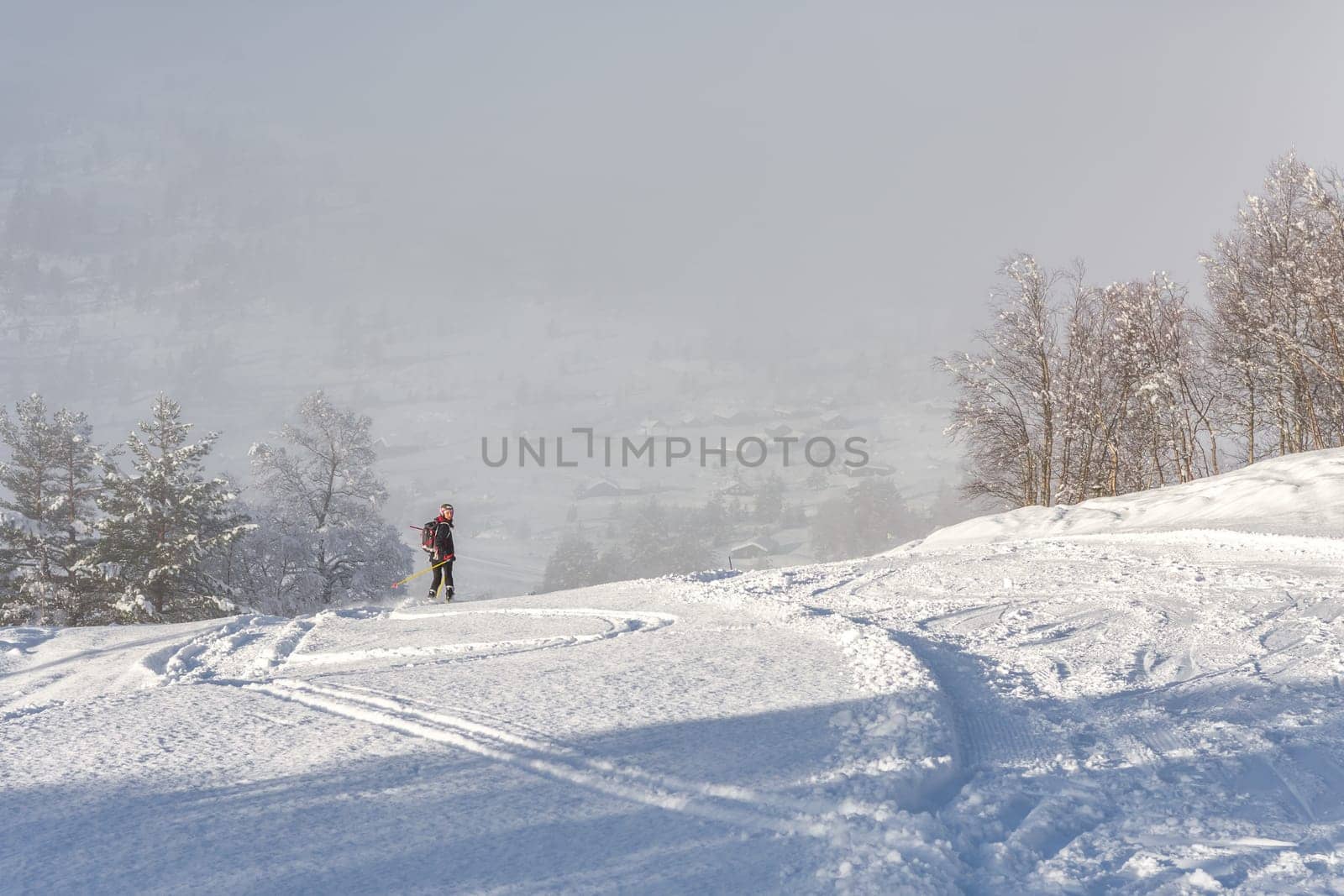 Woman with brown hair, wearing black ski wear and helmet skiing down the top of a mountain, looking back in direction of the camera, rear view, beautiful snow landscape, valley in the distance, fog, horizontal