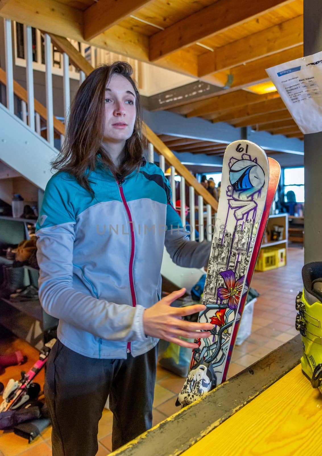Woman with brown hair looking, renting ski equipment, holding her ski while waiting for her helmet and shoes, standing in side a building, staircase behind her, front view, vertical