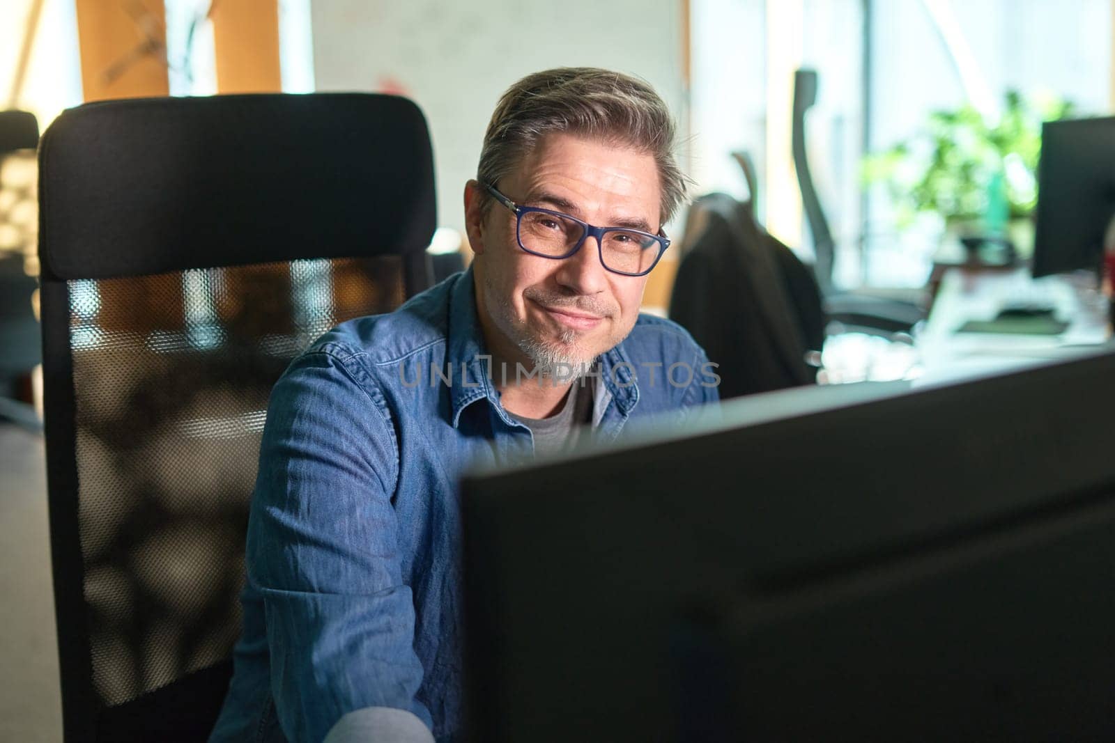 Businessman working with computer in office. Business lifestyle - casual middle aged man sitting at desk in front of screen, smiling, happy.