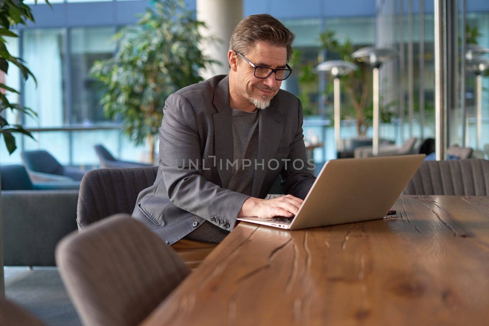 Happy middle age businessman using laptop computer in office. Older casual man working online on terrace of cafe, smiling.