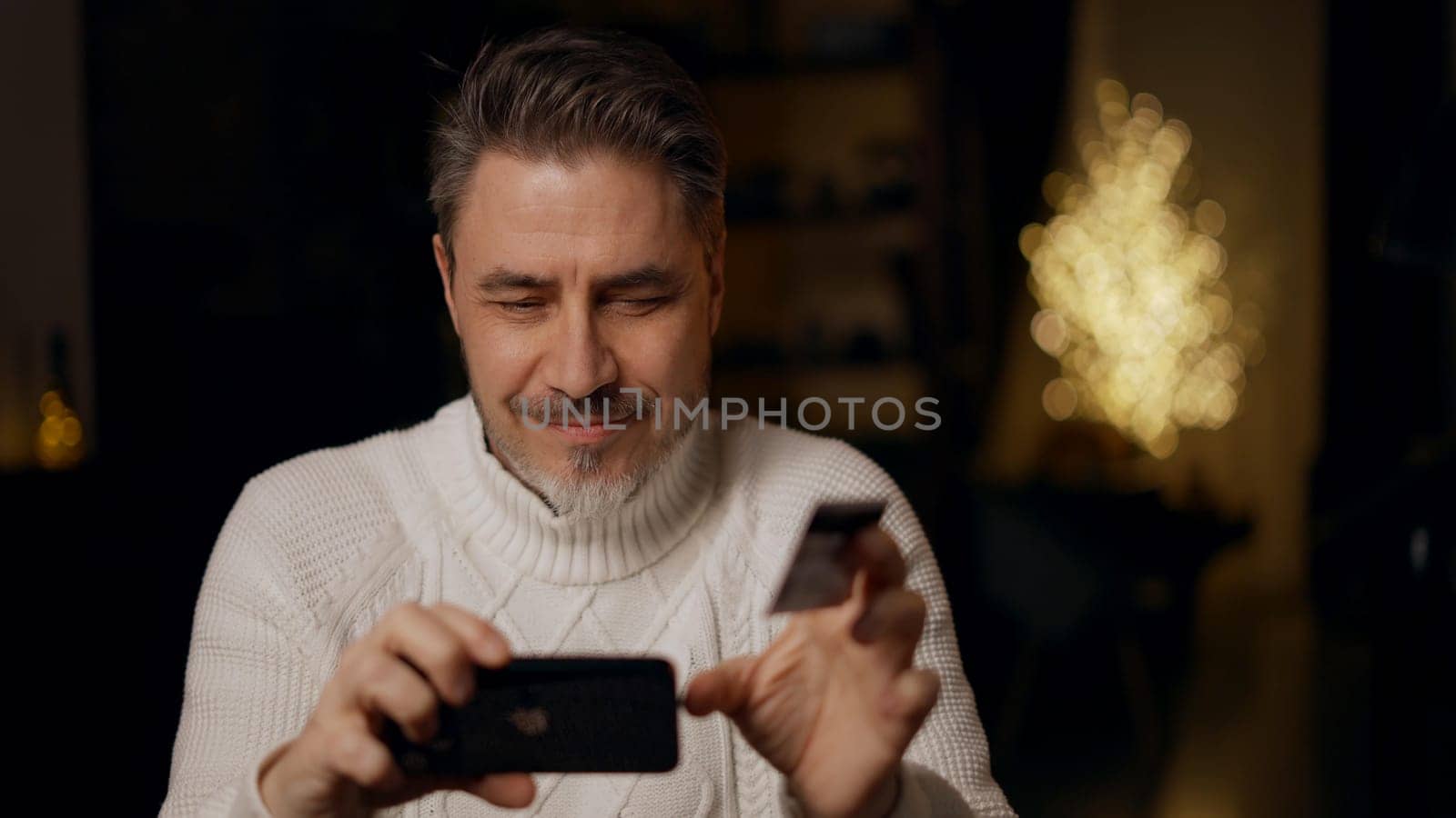 Happy mid adult man at home shopping on the internet using phone paying online with credit card. Winter, Christmas, dark cosy room.