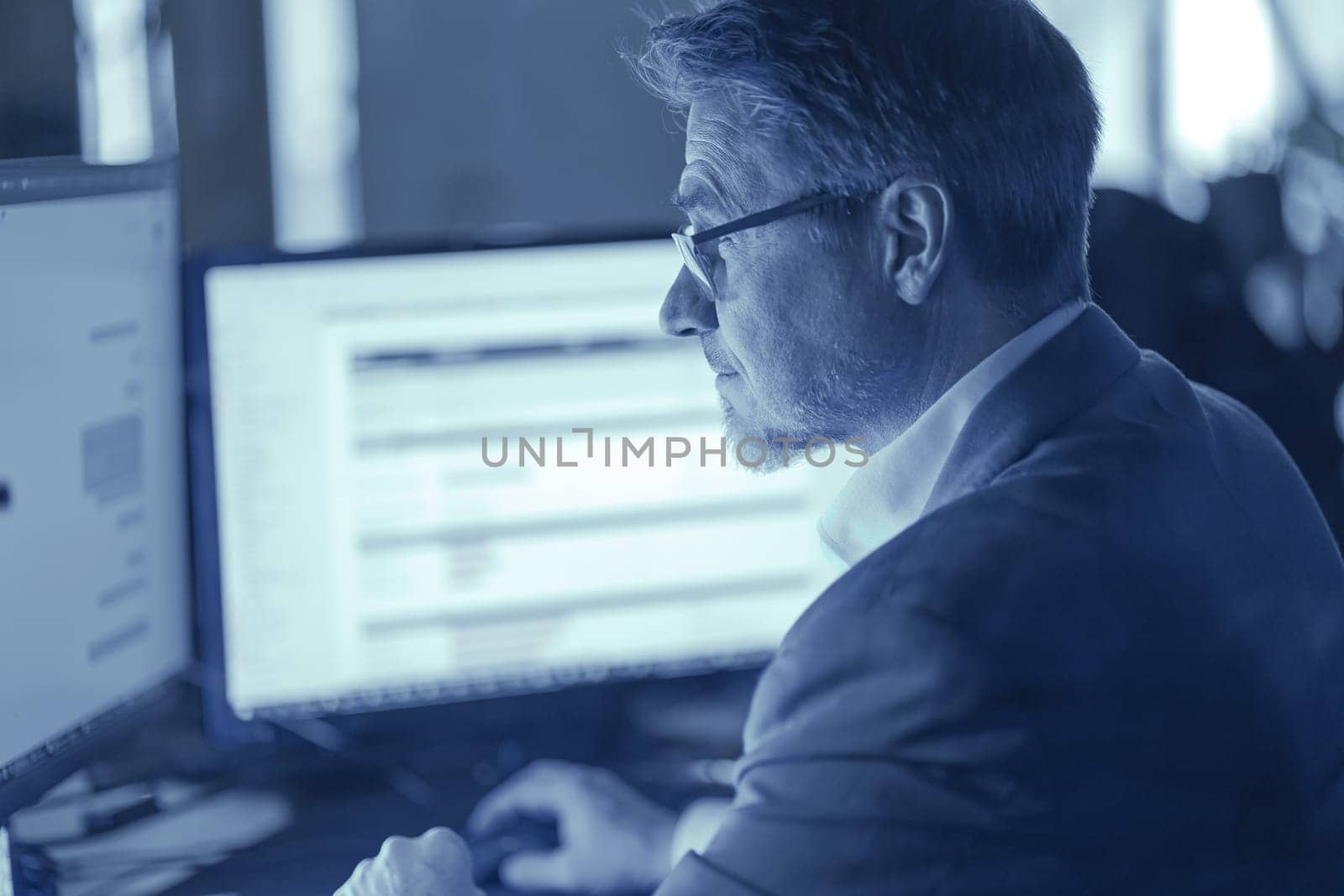 Businessman working with computer at office desk, thinking. Portrait of mid adult man in shirt and jacket, smiling. Bearded, glasses.