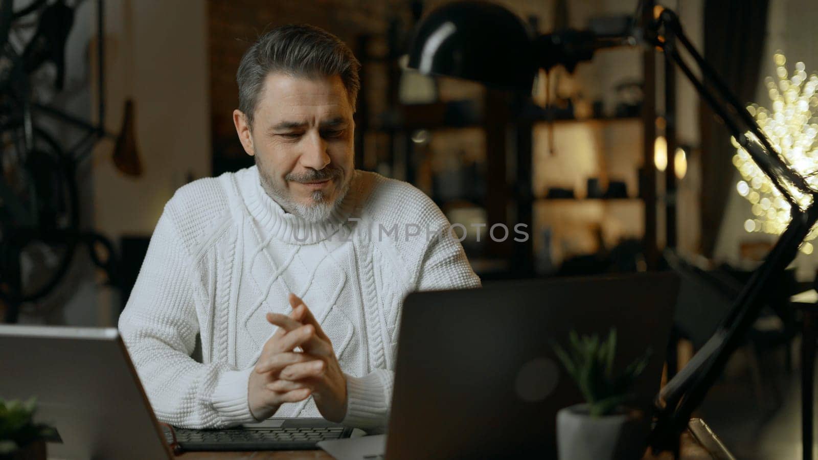 Happy mid adult man working with laptop computer in home office. Winter, Christmas, dark cosy room.