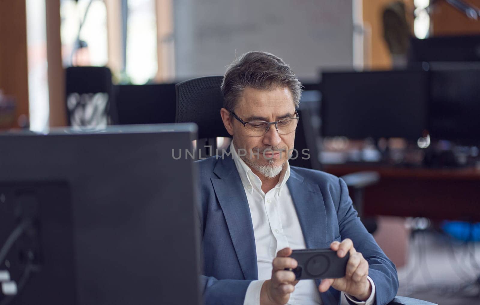 Business portrait - confident businessman sitting at desk working in office, using phone. Happy mid adult man in shirt and jacket, smiling. Bearded, glasses.