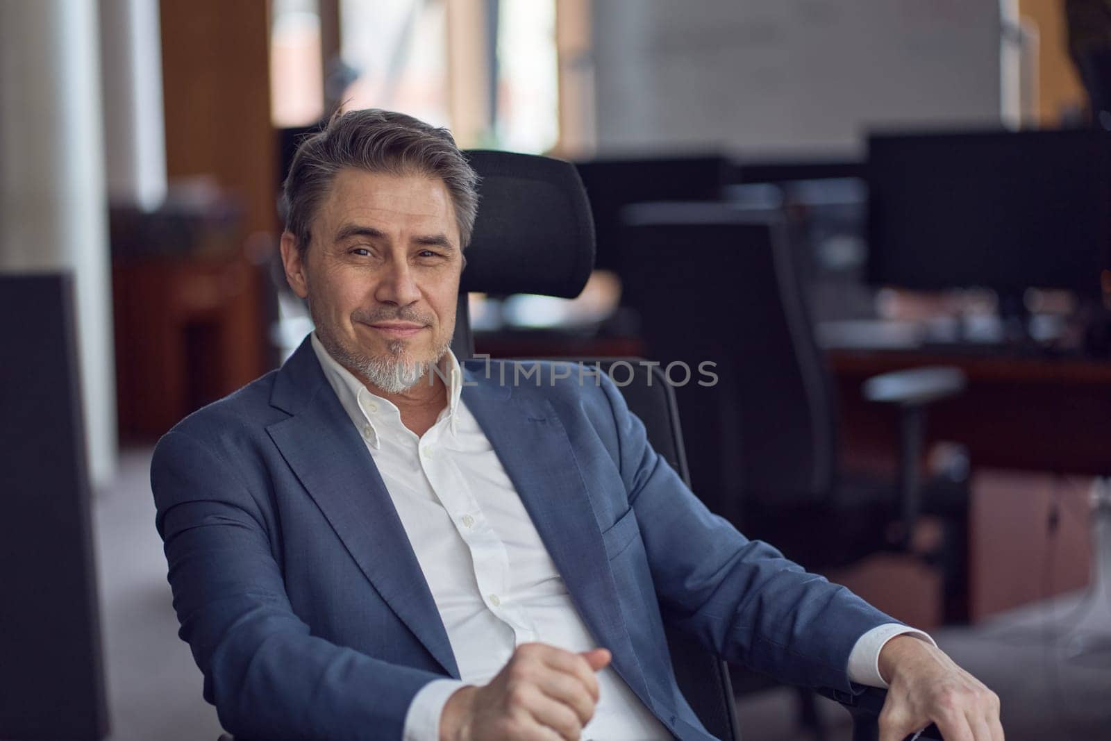 Business portrait - confident businessman sitting at desk in office. Happy mid adult man in shirt and jacket, smiling. Bearded, gray hair.