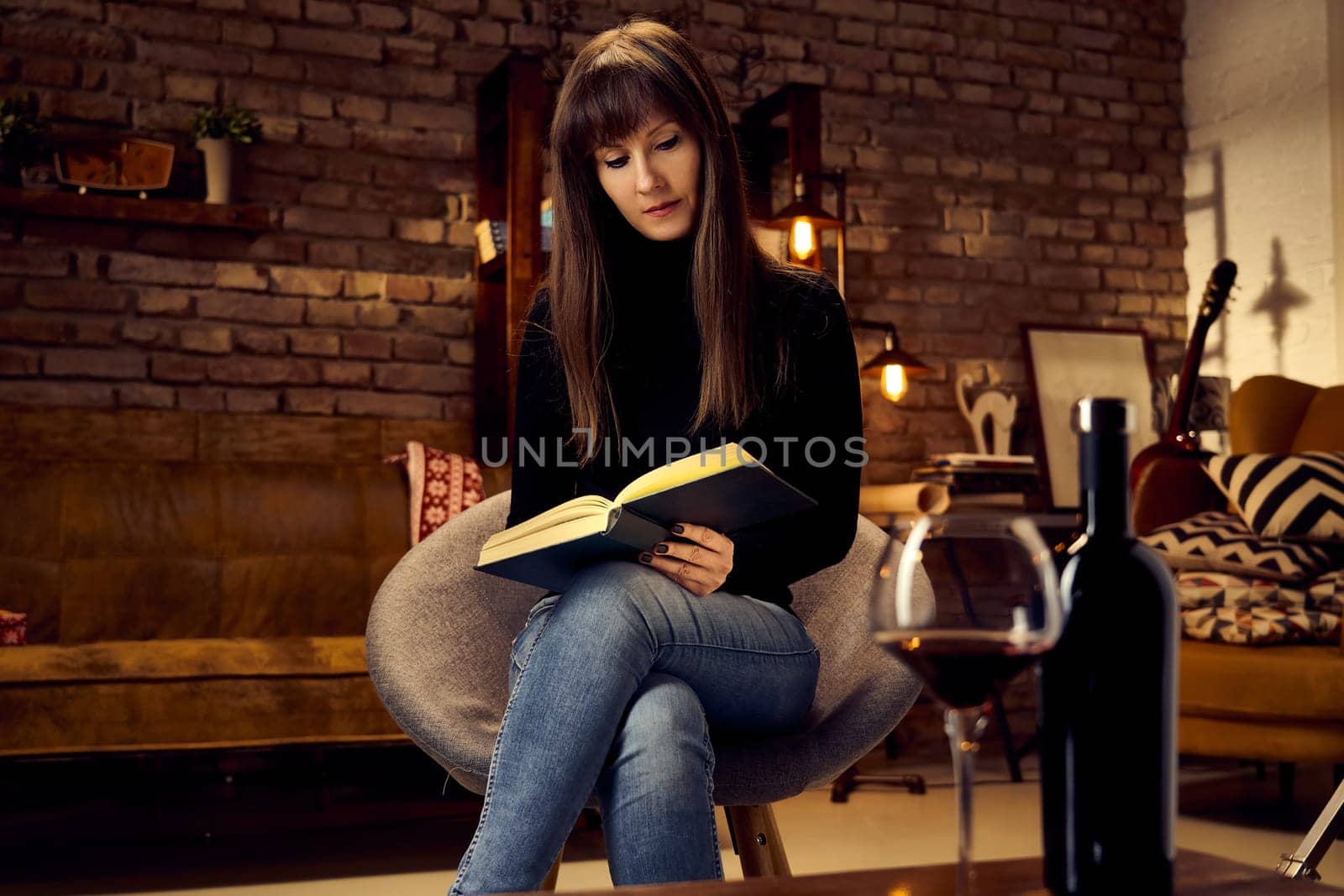 Young adult woman sitting and reading book in cosy dark room. Trendy industrial loft home interior. Autumn or winter mood .