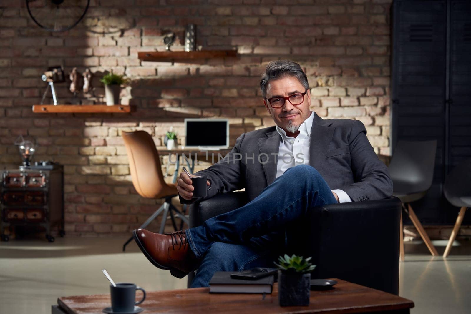 Businessman sitting in armchair in cosy home office, smiling, drinking coffee.