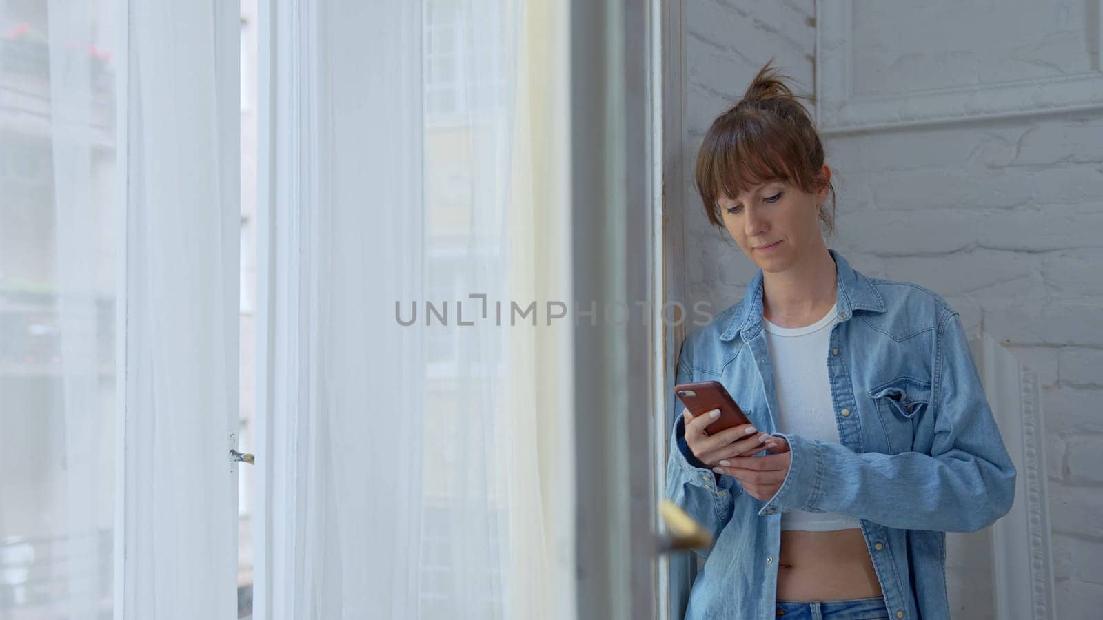 Adult woman standing in window at home daydreaming and checking phone at morning.