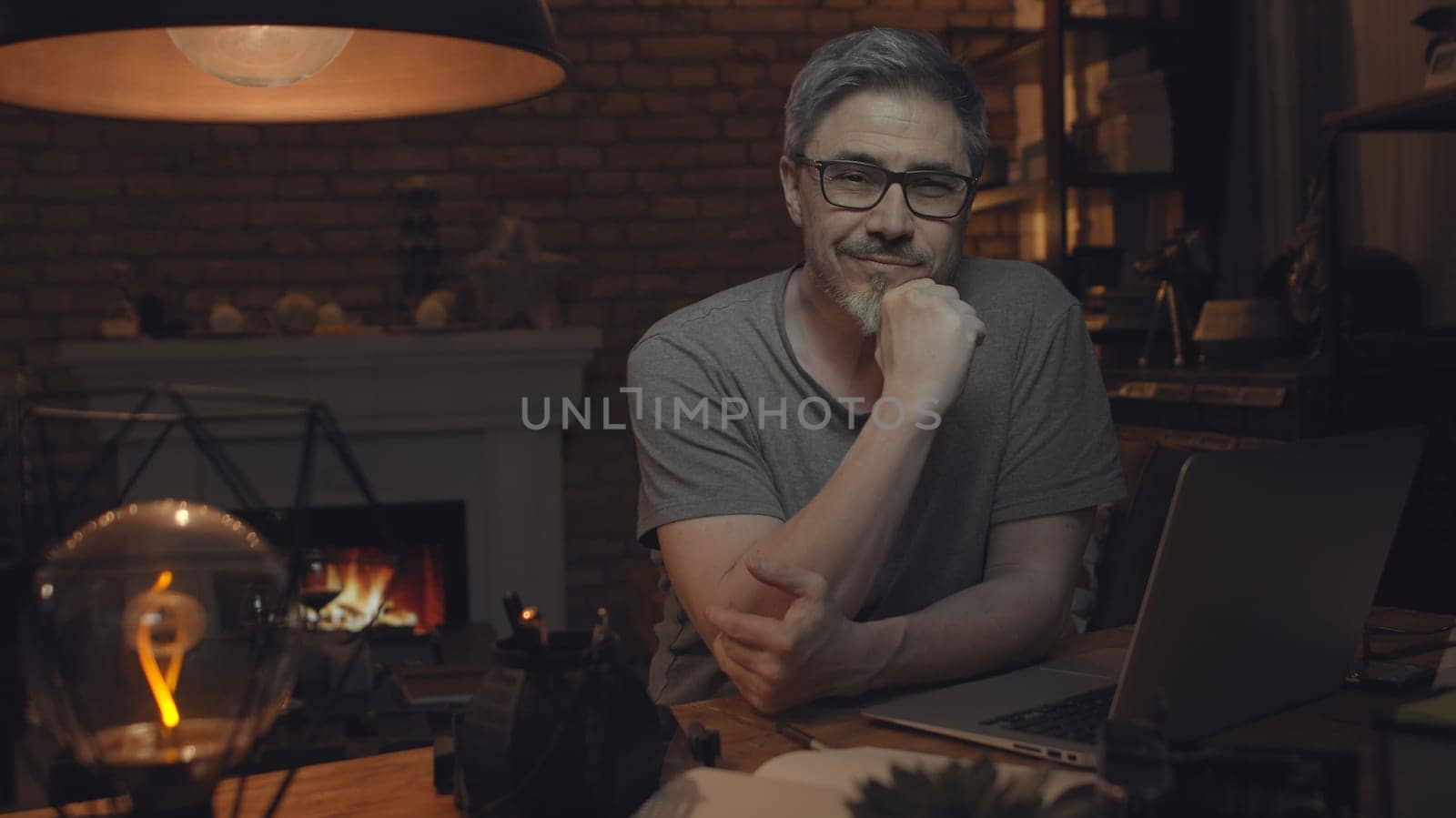 Home office - older white man working from home. Thinking, typing on laptop computer. Sitting at desk in dark living room iin front of fireplace.