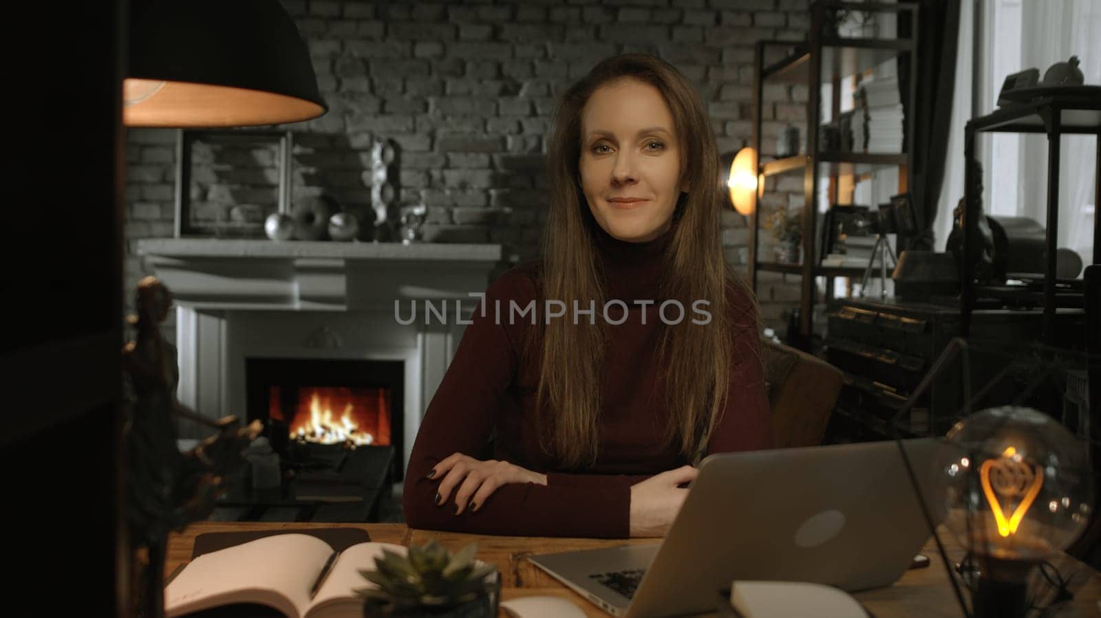 Home office - white woman working from home with laptop computer. Sitting at desk in dark living room in front of fireplace in Winter.