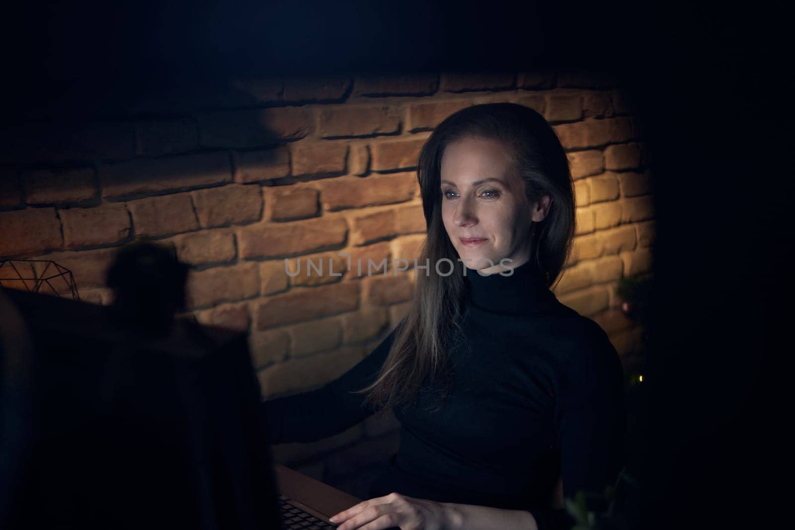 Woman working at desk with computer in dark room at home, smiling.