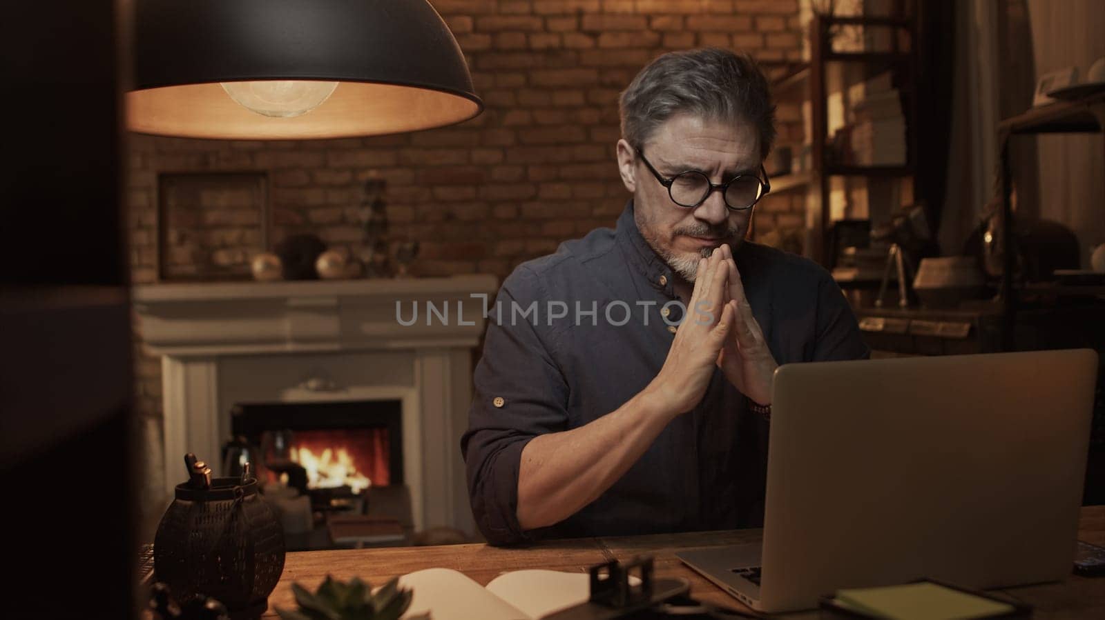 Home office - older white man working from home. Thinking, typing on laptop computer. Sitting at desk in dark living room iin front of fireplace.