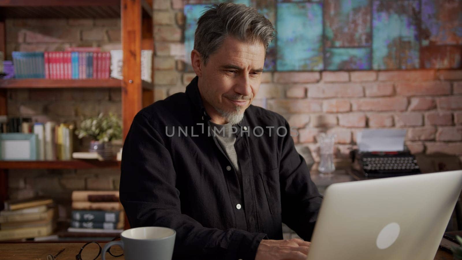 Mature man working from home, sitting at desk in home office using laptop, thinking, browsing internet, teleworking online.