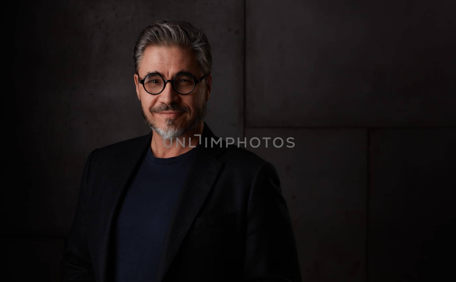 Portrait of happy older man in 50s with gray hair and glasses against dark background. Wearing jacket, smiling, copy space.