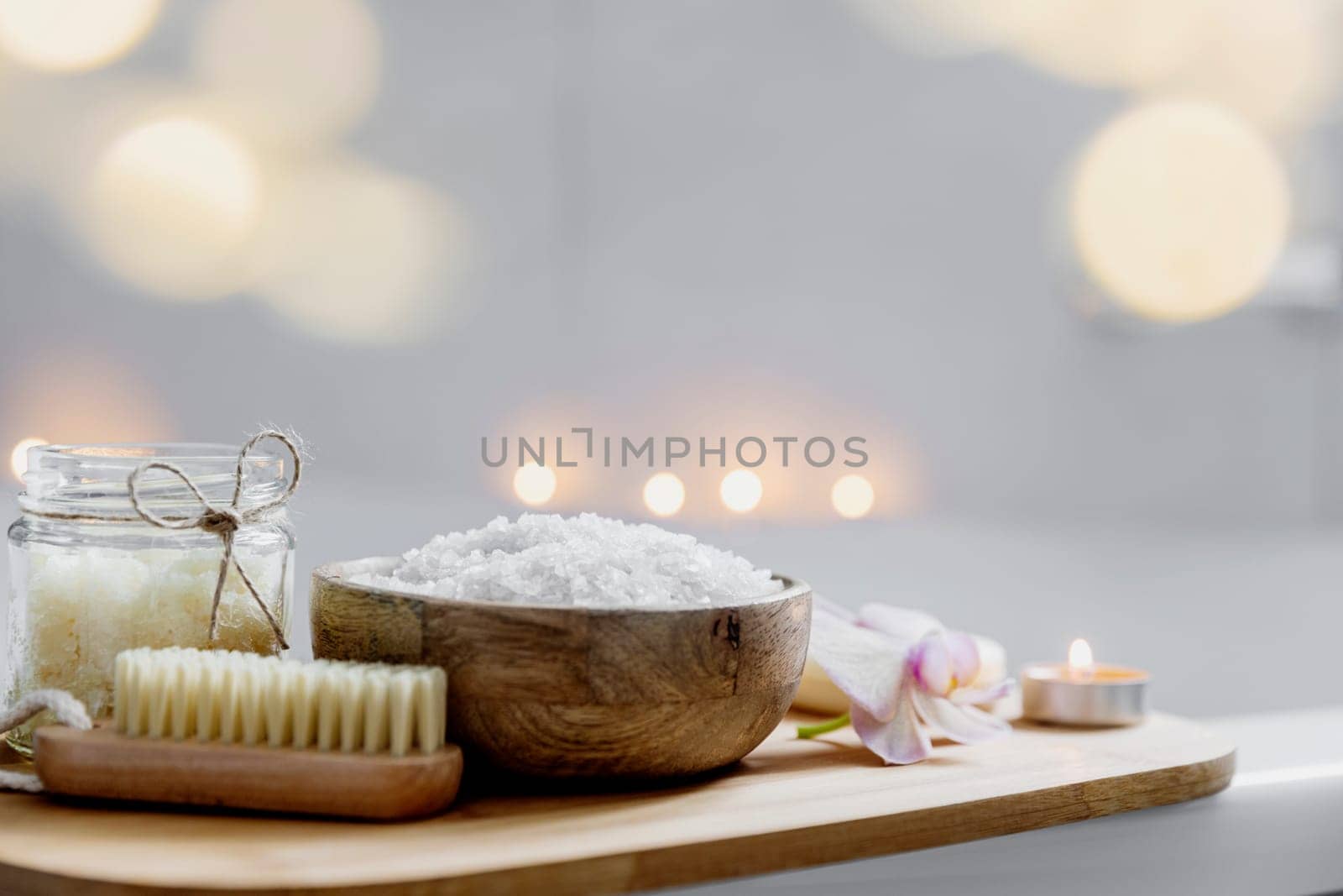 Preparation for hotel spa treatment, home bath procedure. White washbasin in bathroom, accessories on tray. Burning candles, soap, foot brush, towel, glass bottle with sea salt, orchid flower