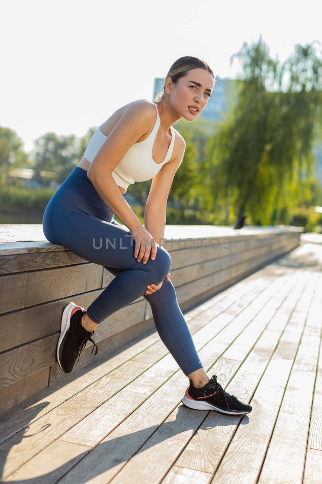 Young sportswoman sitting with knee pain in city park. Woman massaging stretching, trauma injury while jogging outdoors. Fitness female sprain severe pain stretch pull. Leg muscle cramp calf sport.