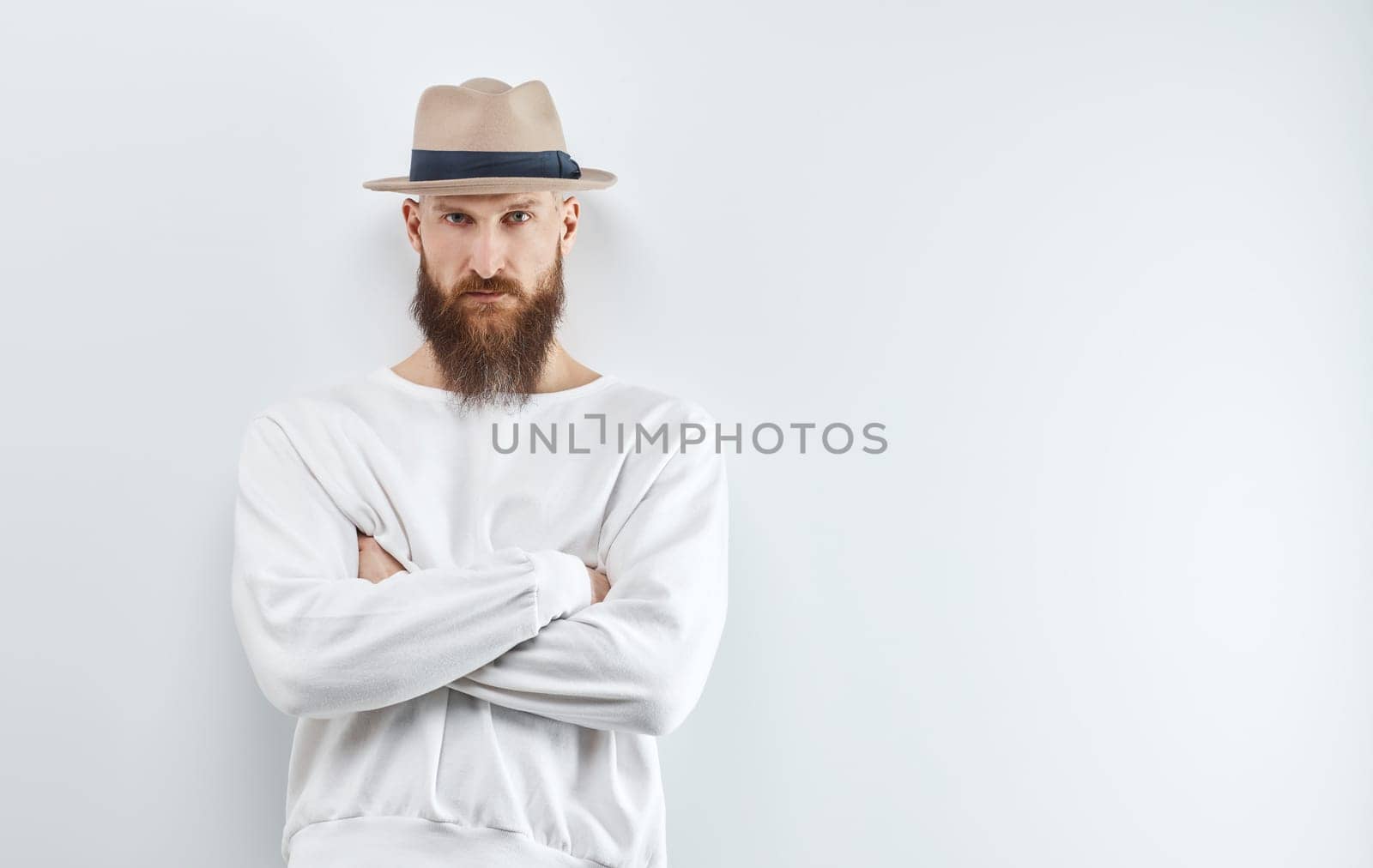 Serious bearded man ib hat standing against white wall.