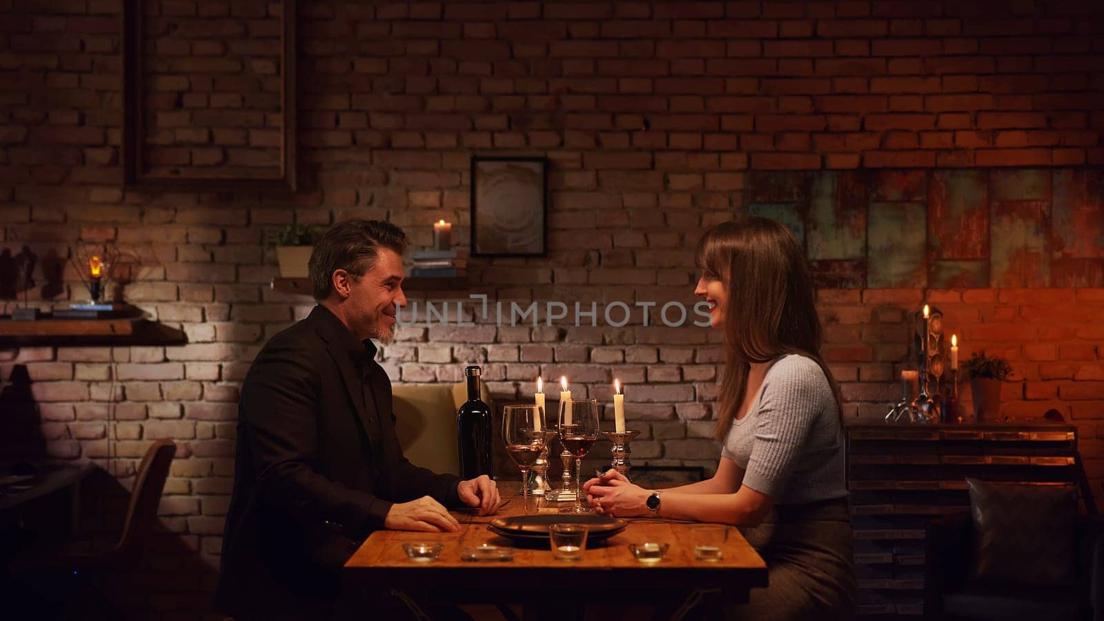Elegant couple having romantic dinner at home, sitting at table in living room drinking red wine, talking. Stay at home concept.