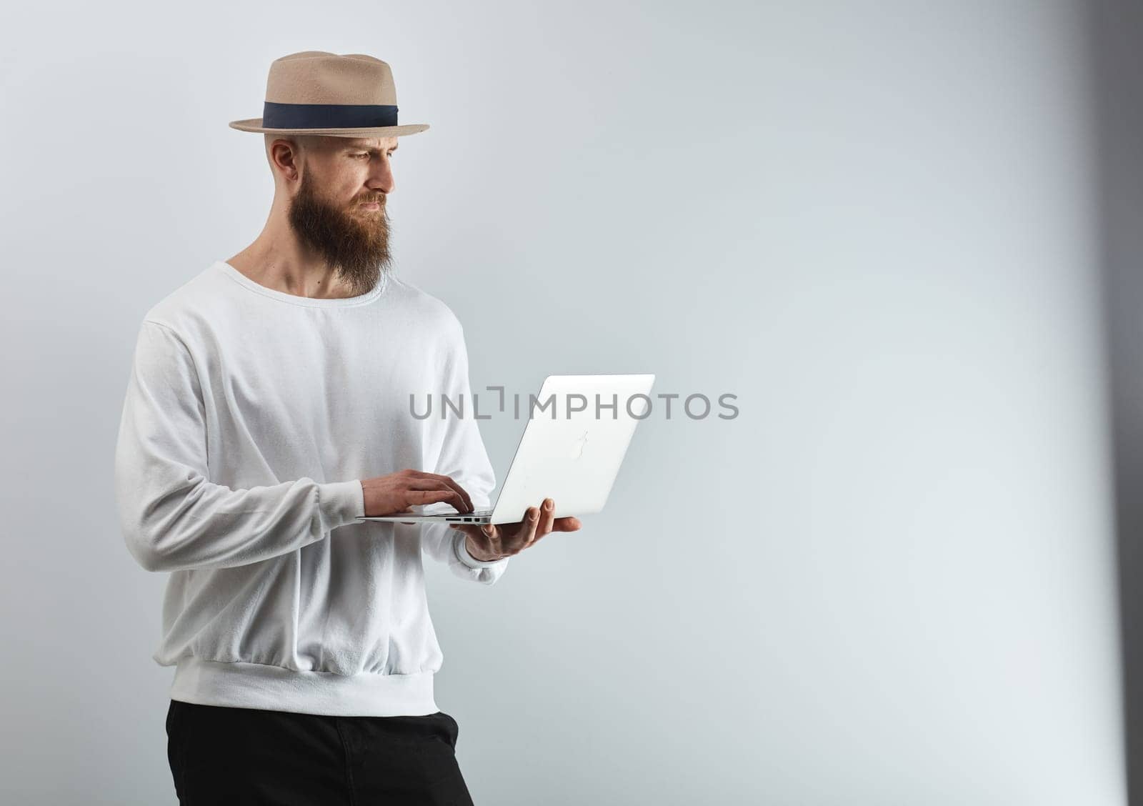 Serious bearded man in hat standing against white wall holding laptop computer.