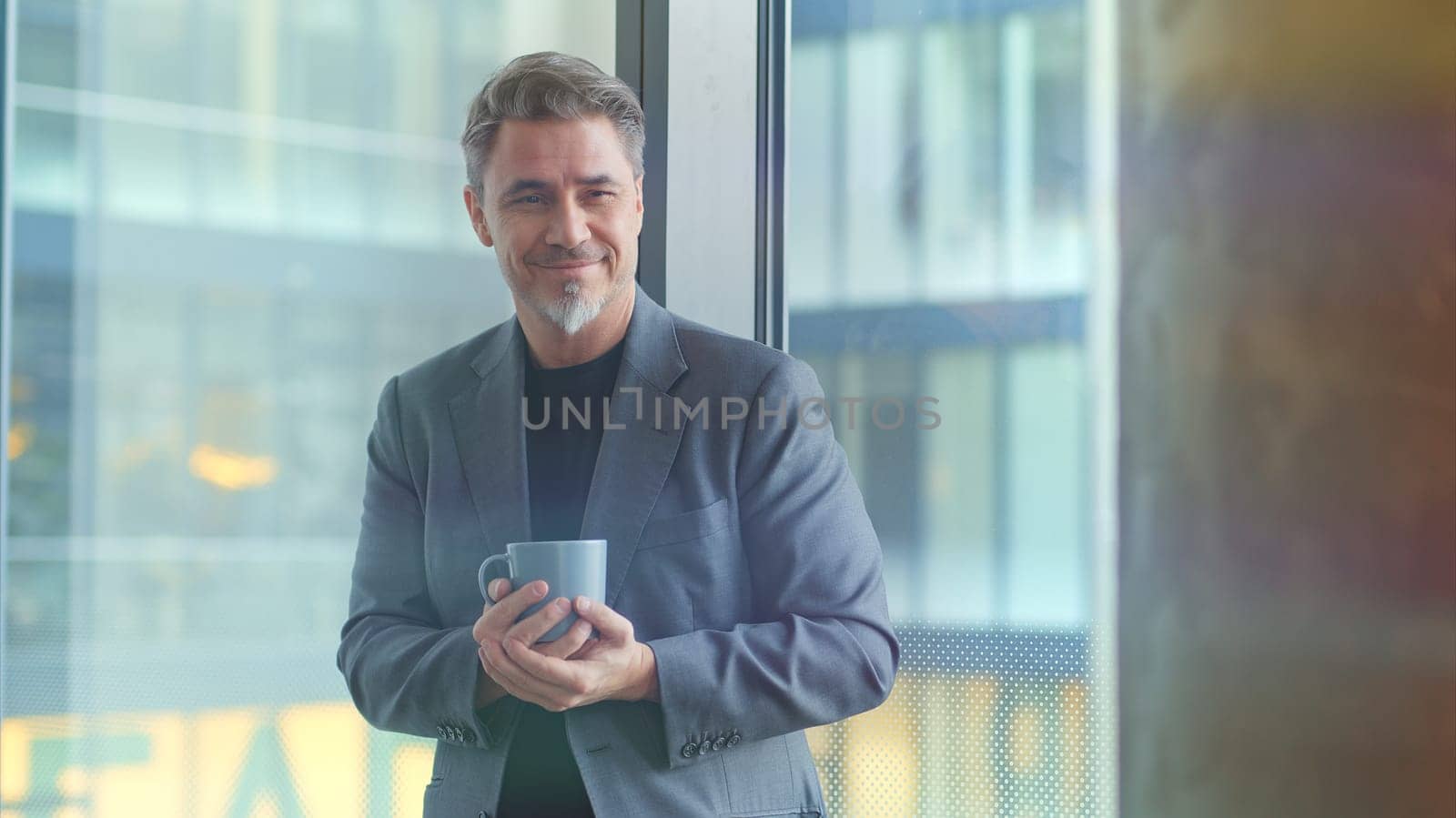 Happy older businessman taking break in office drinking coffee standing in window and daydreaming, thinking.