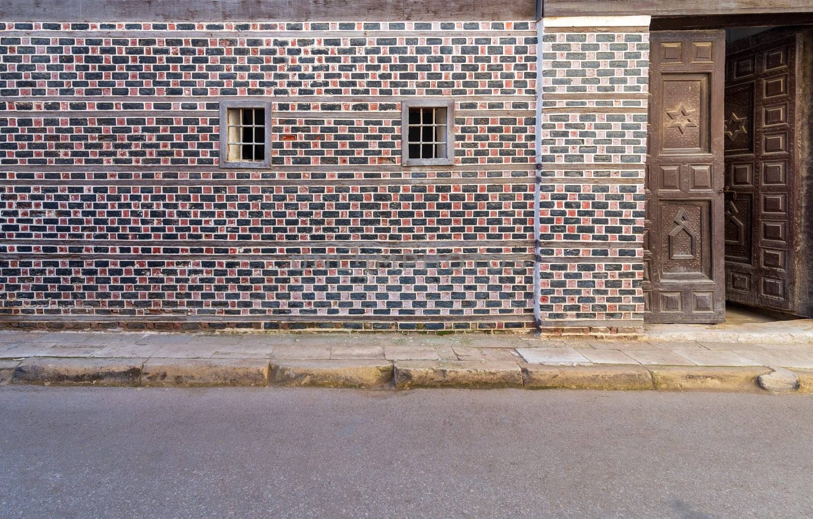 Wall with black and red bricks with white seam and two small adjacent windows with metal bars on cobblestone road, and wooden door with engraved Star of David